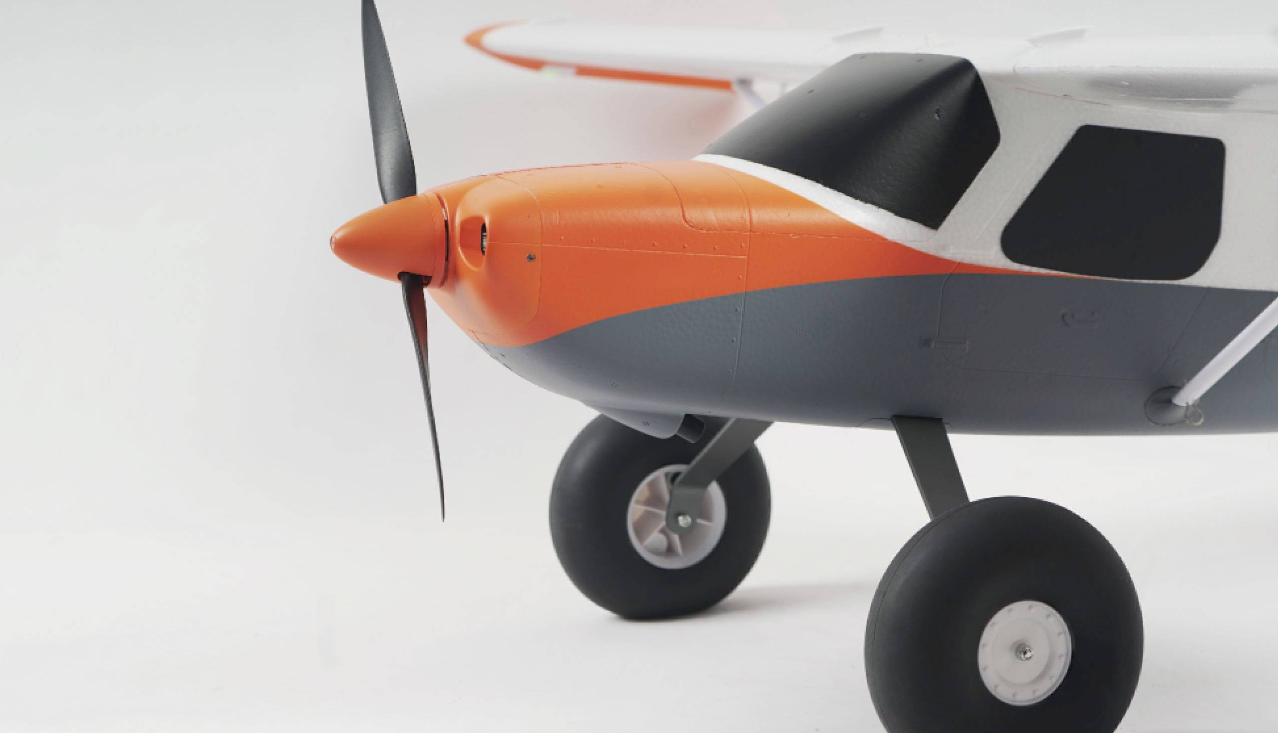 Close-up van een modelvliegtuig met een oranje-grijze neus en een propeller. Het vliegtuig heeft grote wielen en staat op een witte achtergrond.