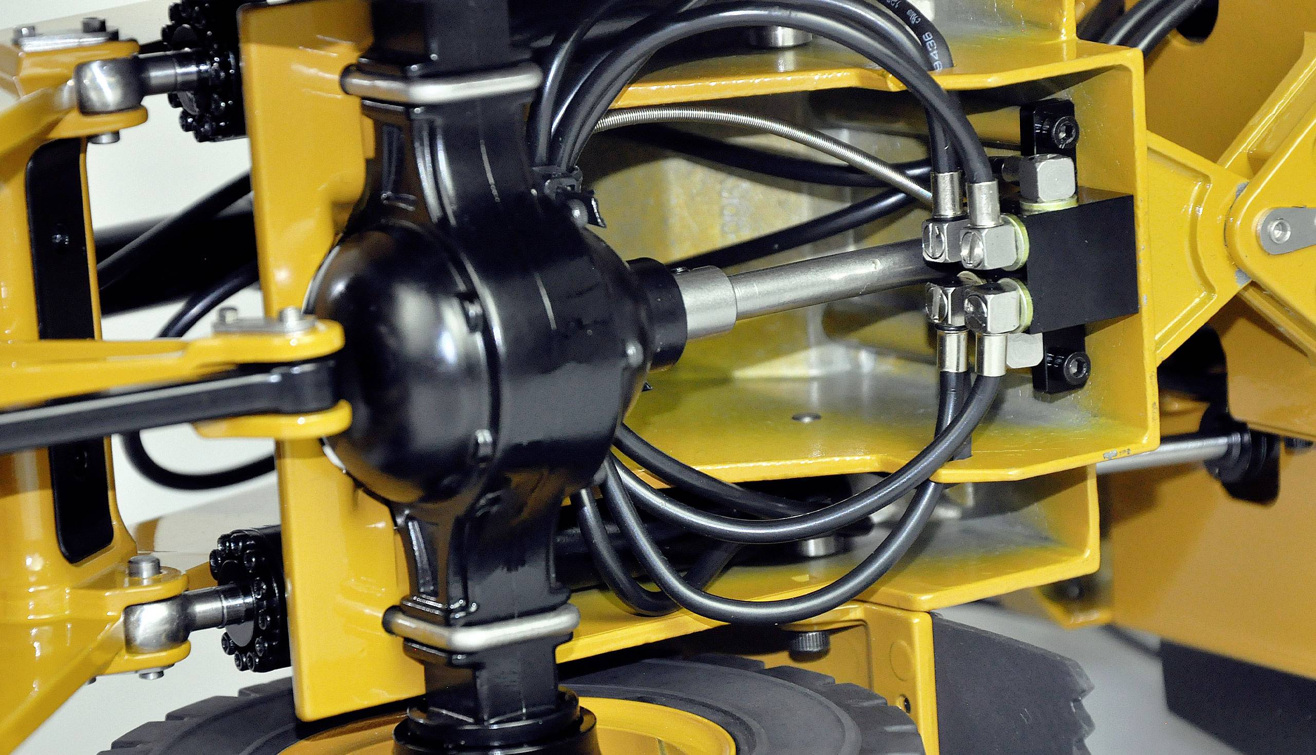 Close-up van een geel mechanisch apparaat met zwarte hydraulische leidingen en metalen verbindingen. Details van buizen en schroeven zijn zichtbaar.