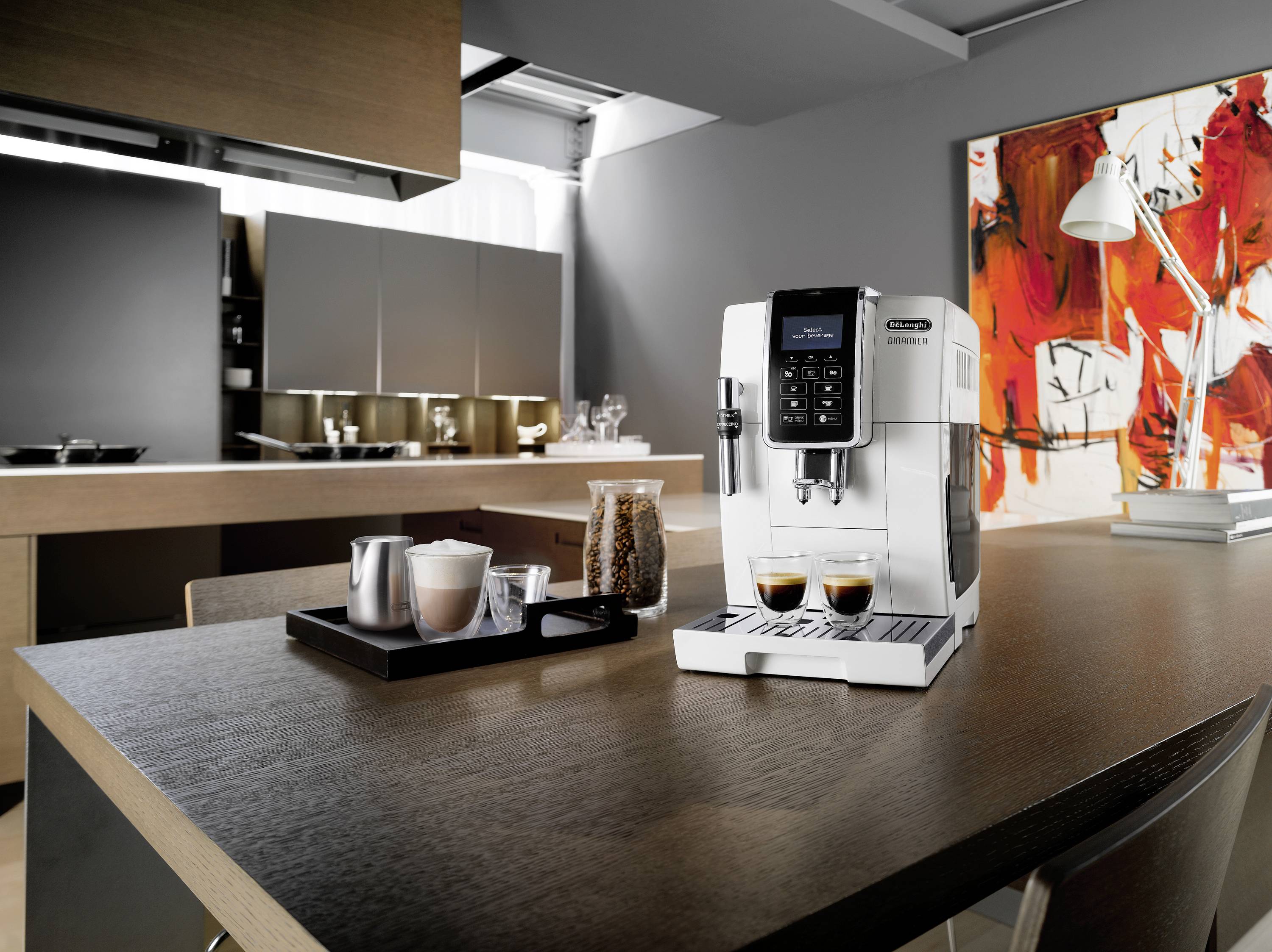 Een koffiemachine staat op een keukentafel. Op de achtergrond zijn moderne keukenkastjes en een kleurrijk schilderij aan de muur te zien.