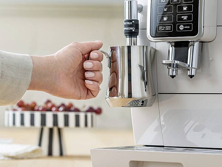Een hand houdt een zilveren melkschuimkan onder de uitloop van een witte koffiemachine. Op de achtergrond staat een schaal met druiven.