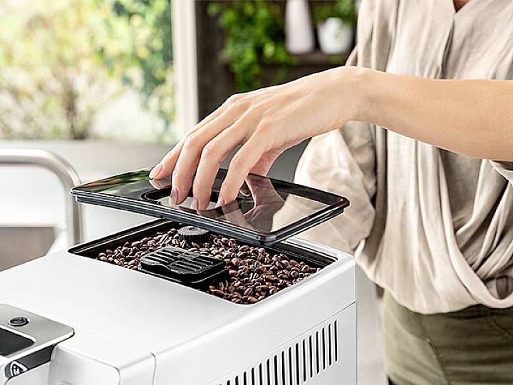 Een persoon opent de bonenhouder van een koffiemachine om bonen bij te vullen. De scène speelt zich af in een keuken.