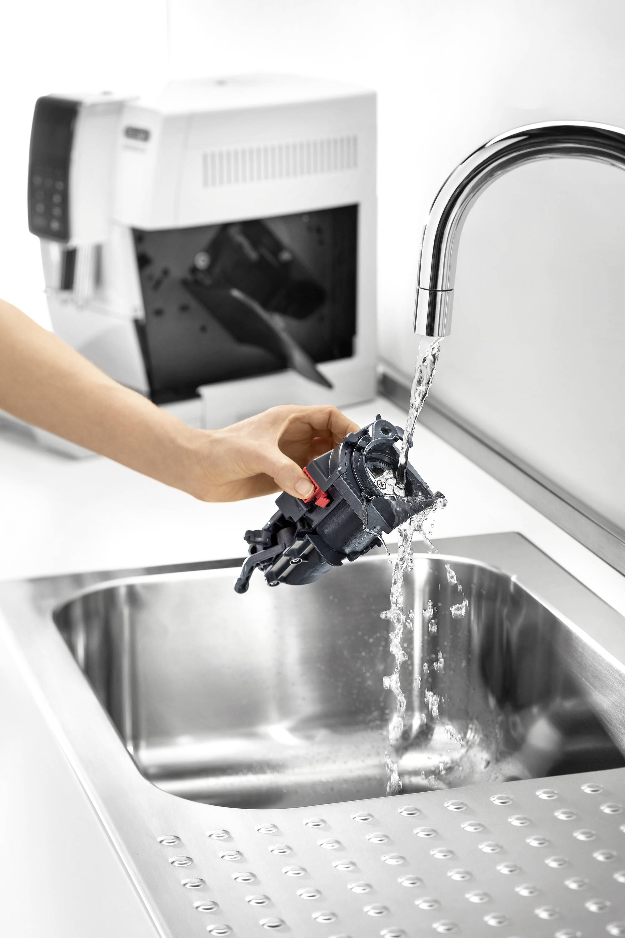 Een persoon reinigt een onderdeel van een koffiemachine onder stromend water in een roestvrijstalen gootsteen. Op de achtergrond staat een koffiemachine.