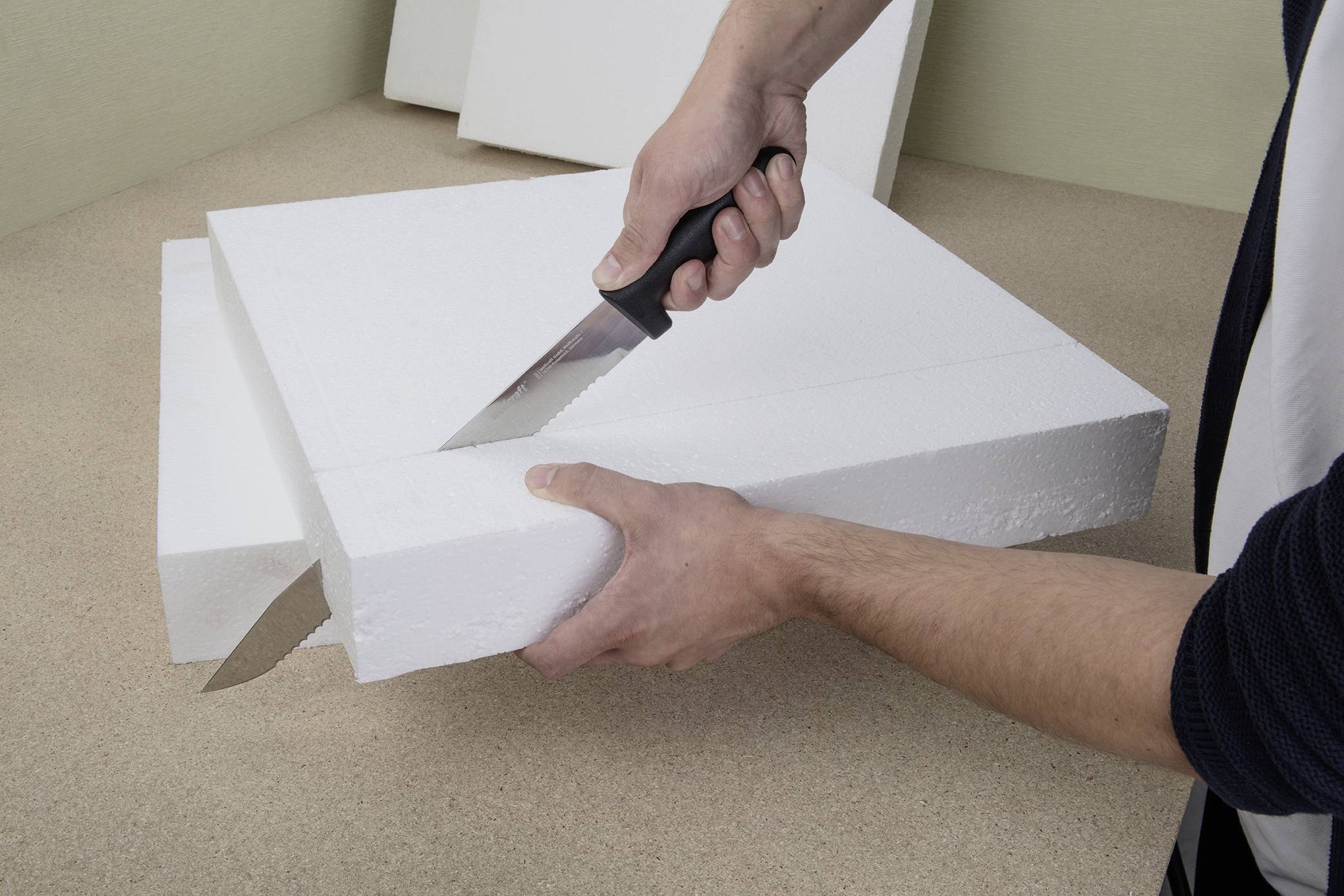 'Een persoon snijdt een grote piepschuimblok met een mes. De ondergrond is een tafel of werkoppervlak.'