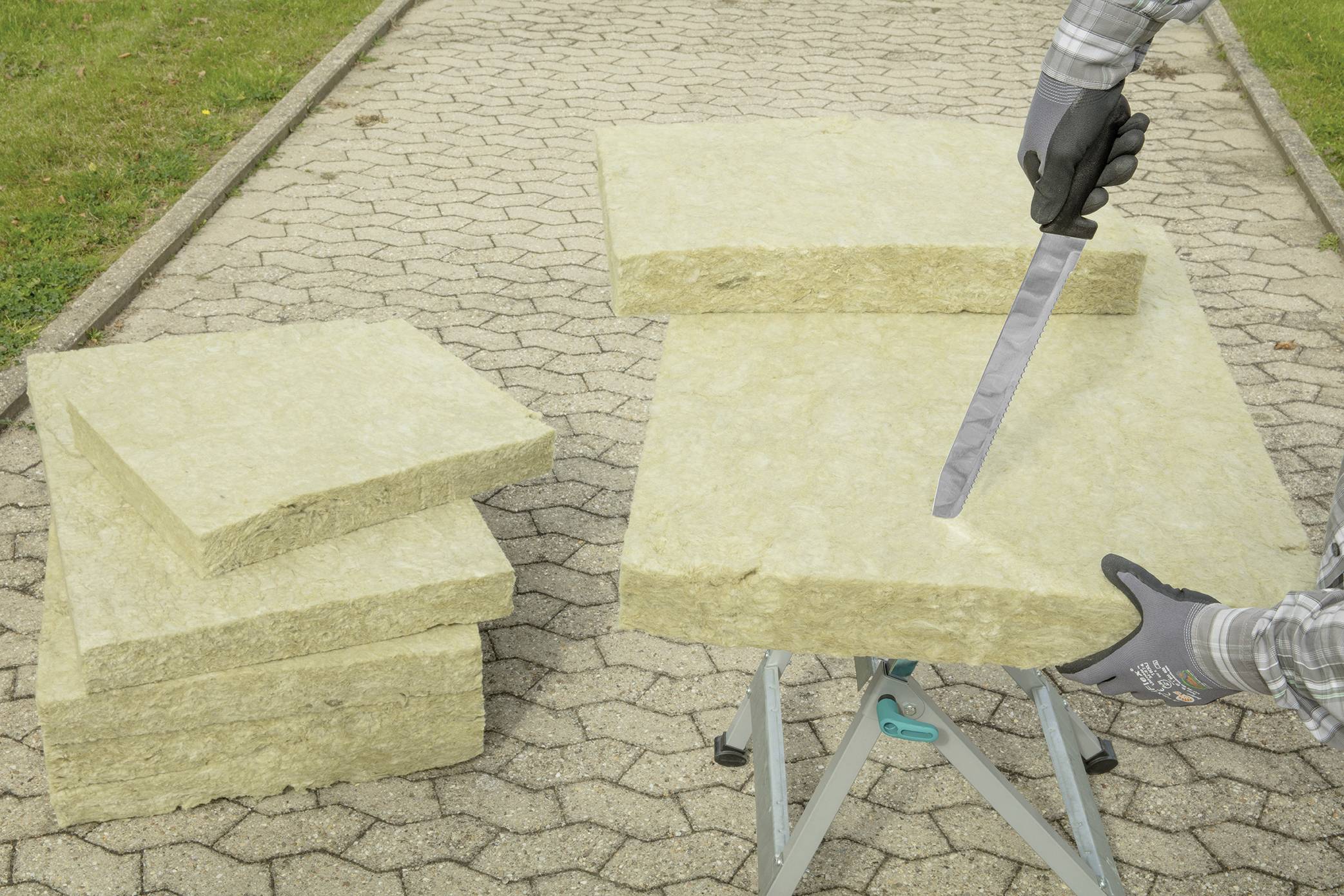 Een persoon snijdt mineraalwol-isolatieplaten met een mes op een schraag buiten. Verdere platen liggen gestapeld ernaast.