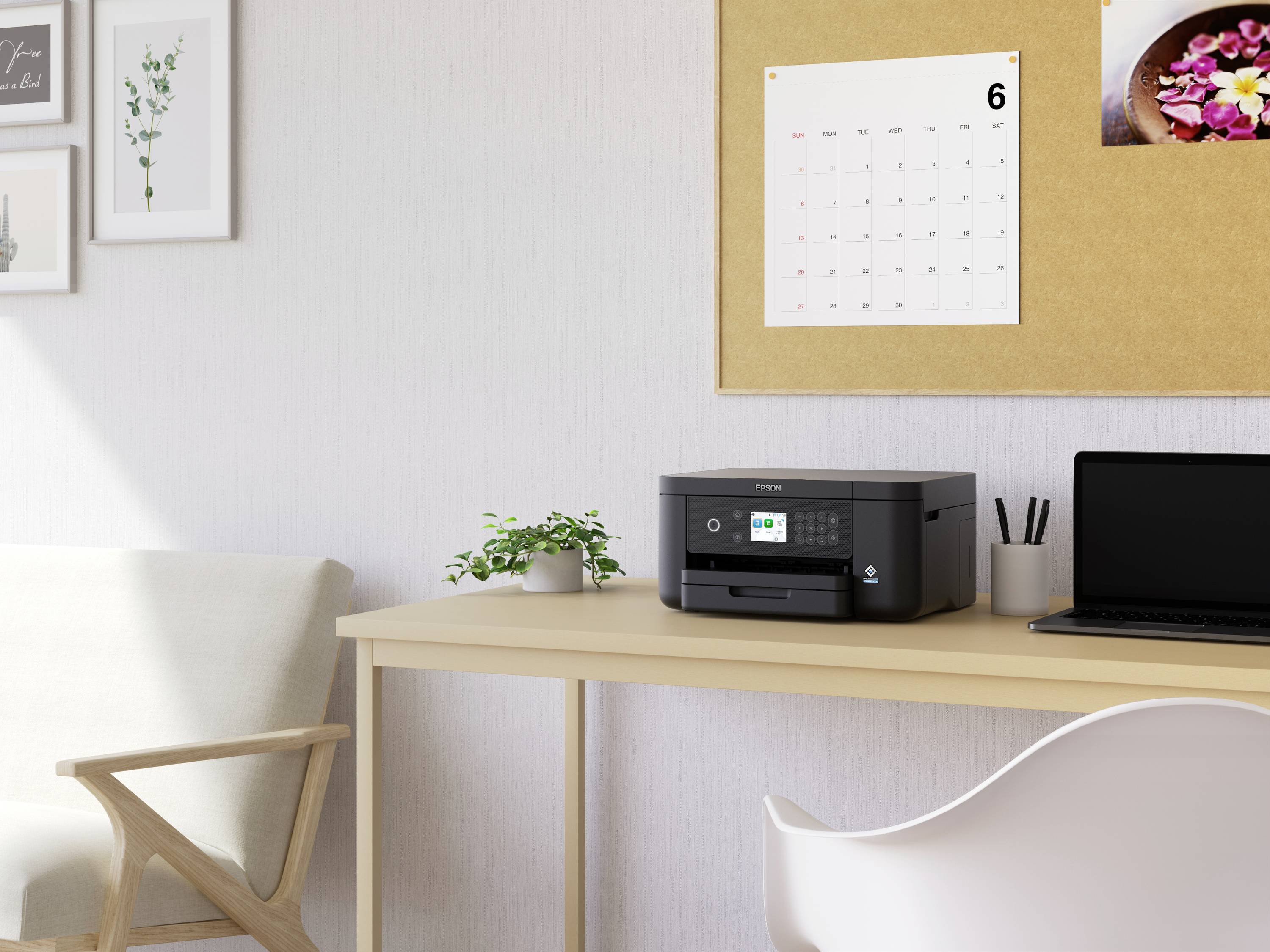 Een printer staat op een houtkleurig bureau naast een laptop. Aan de muur hangt een kalender met de maand juni.