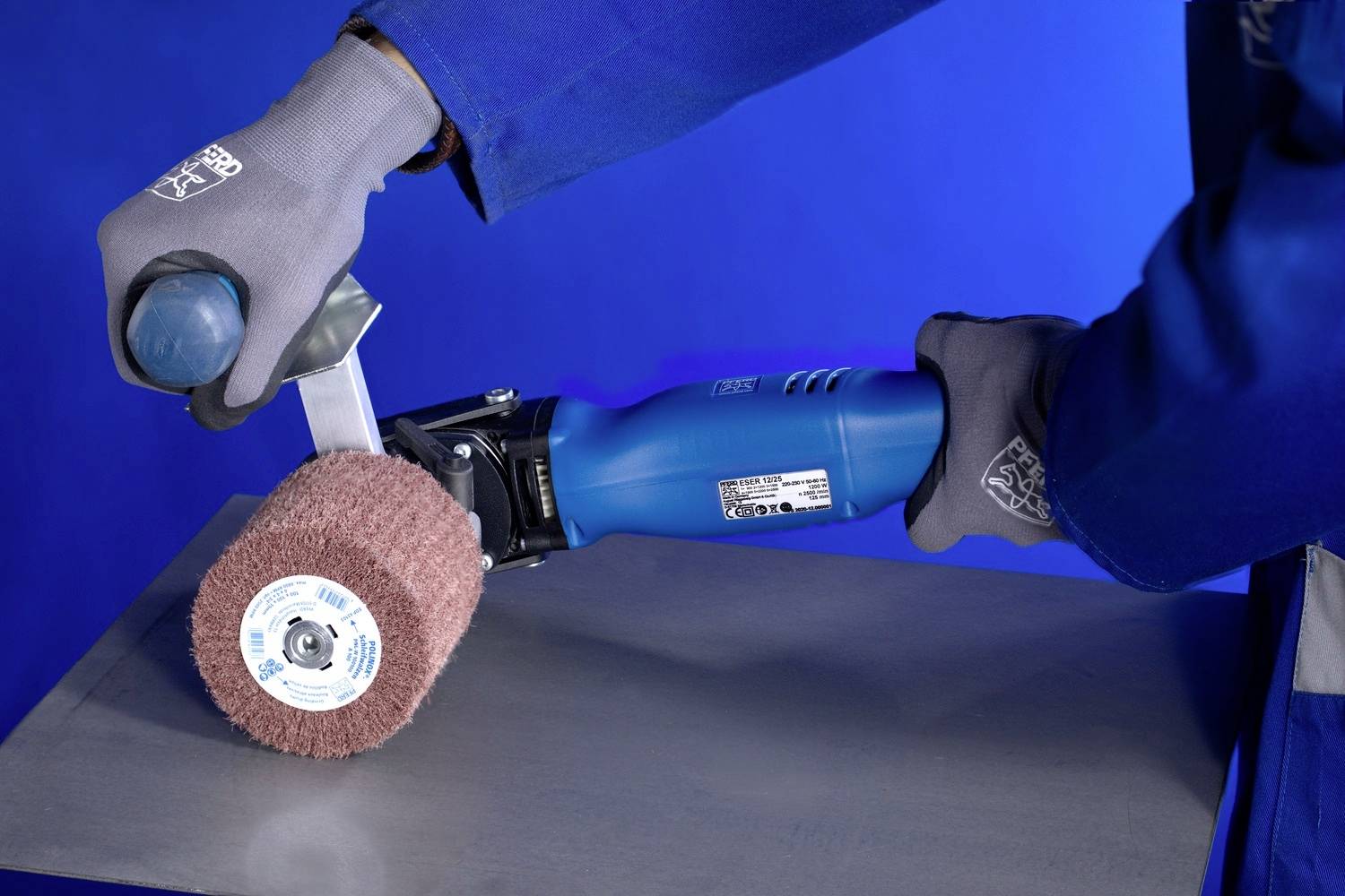 Een persoon slijpt een metalen plaat met een elektrische slijpmachine in een blauwe omgeving. Werkkleding en handschoenen zijn zichtbaar.