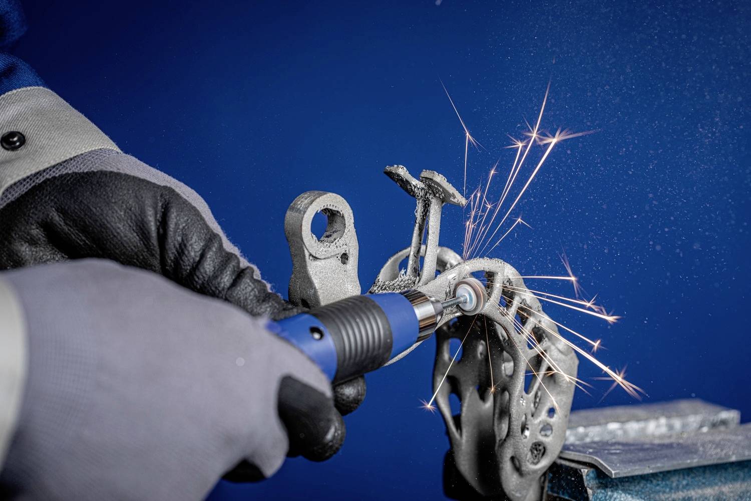 Een persoon slijpt metaal met een elektrisch gereedschap, vonken spatten. Hij draagt handschoenen en veiligheidskleding voor een blauwe achtergrond.