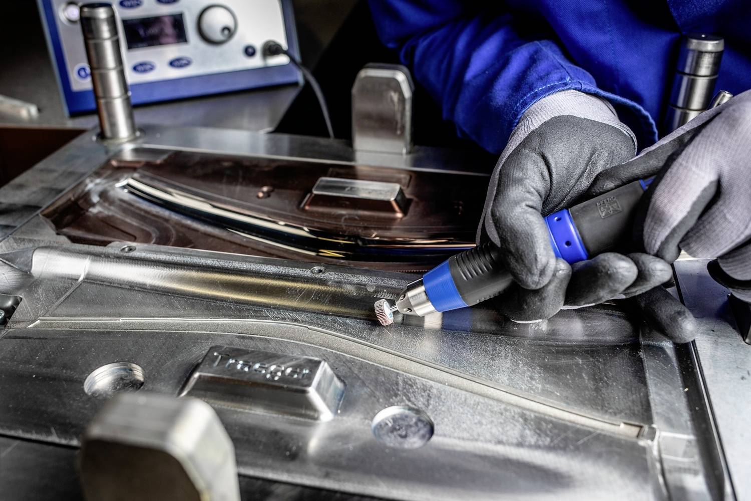 Een arbeider polijst nauwkeurig een metalen oppervlak met een handgereedschap in een werkplaats. Op de achtergrond een meetinstrument.