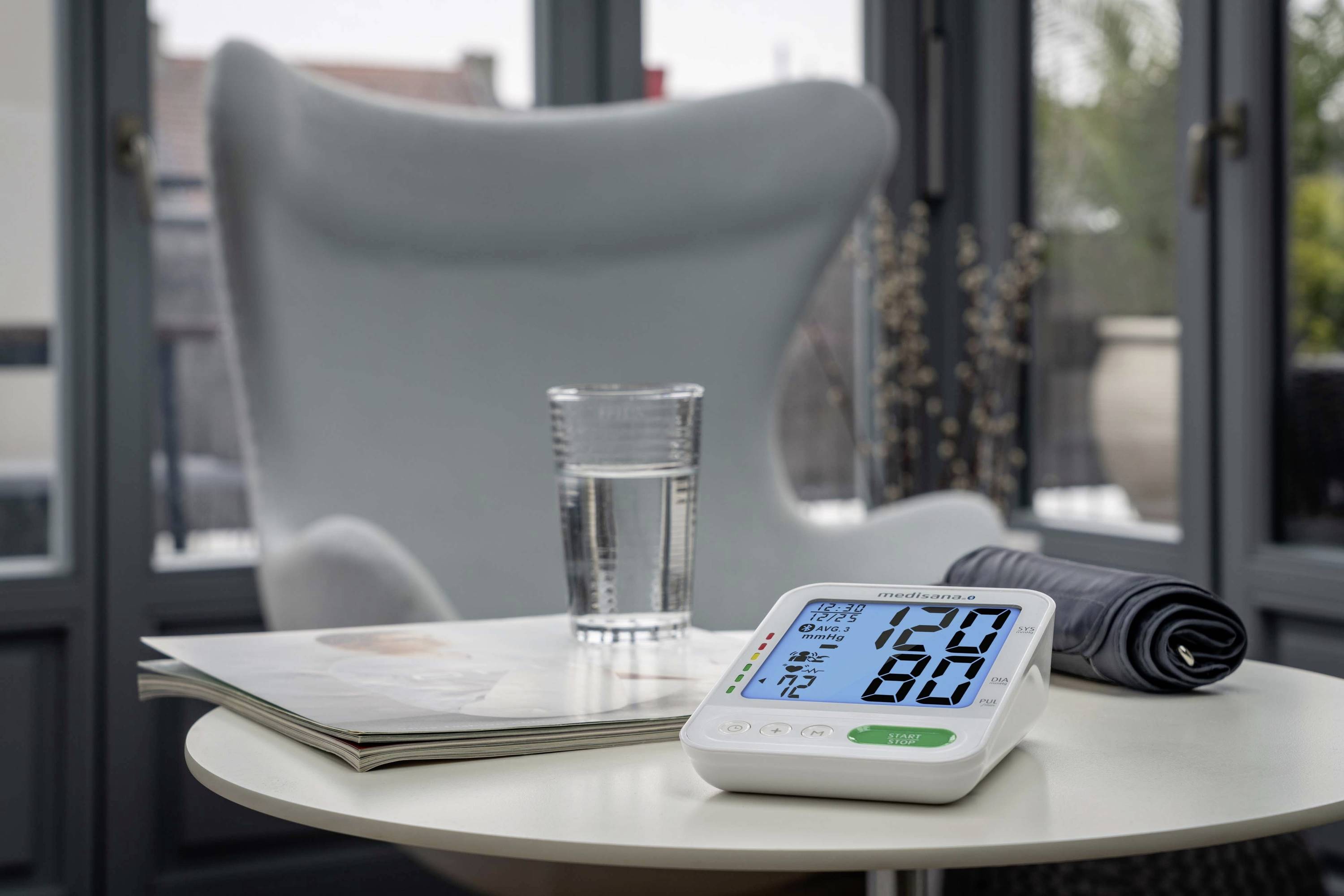 Een digitaal bloeddrukmeetapparaat toont de waarde 120/80. Het staat op een tafel naast een glas water en een tijdschrift.