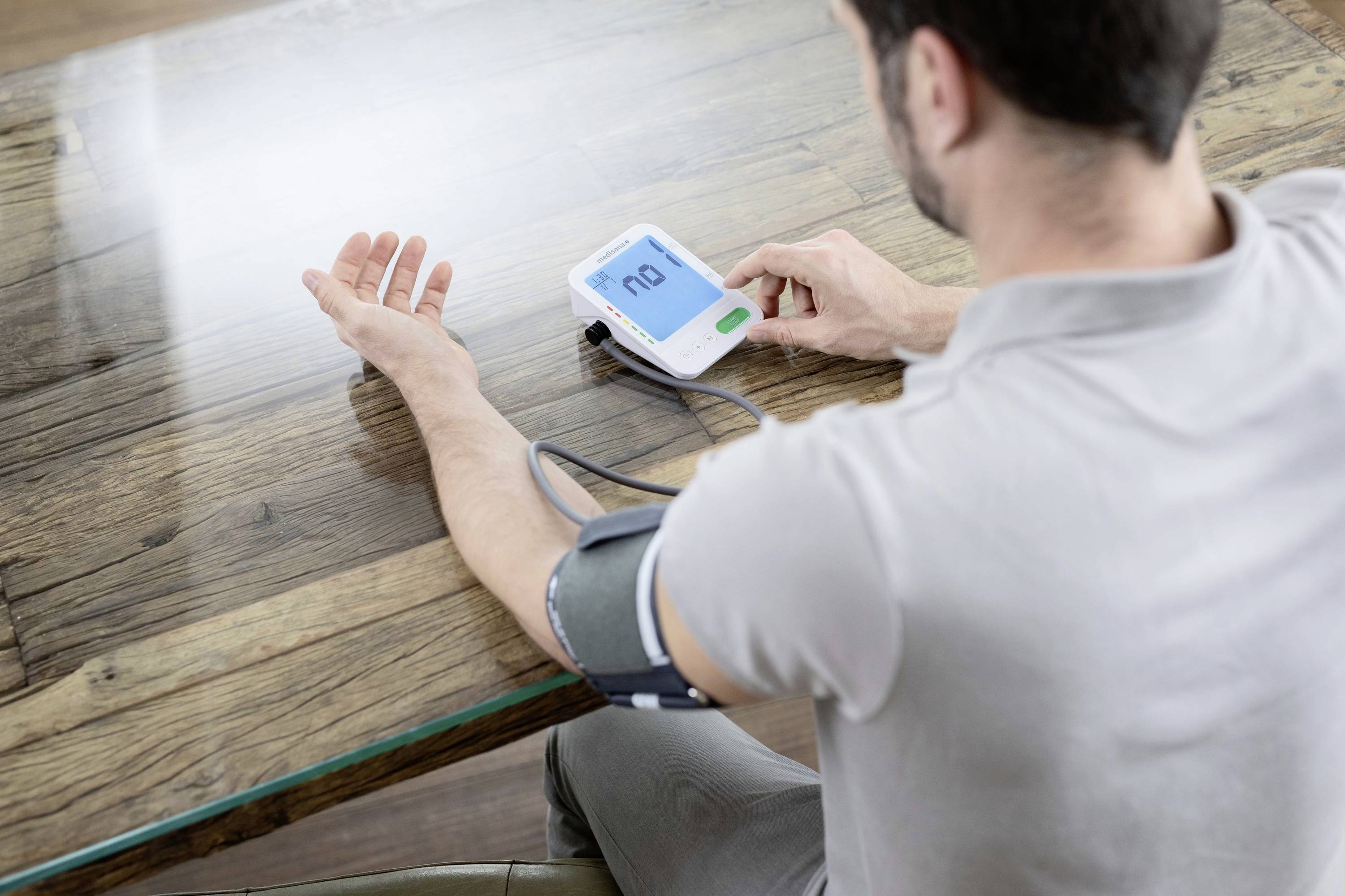 Een man meet zijn bloeddruk met een digitaal apparaat aan zijn linkerarm. Hij zit aan een houten tafel.