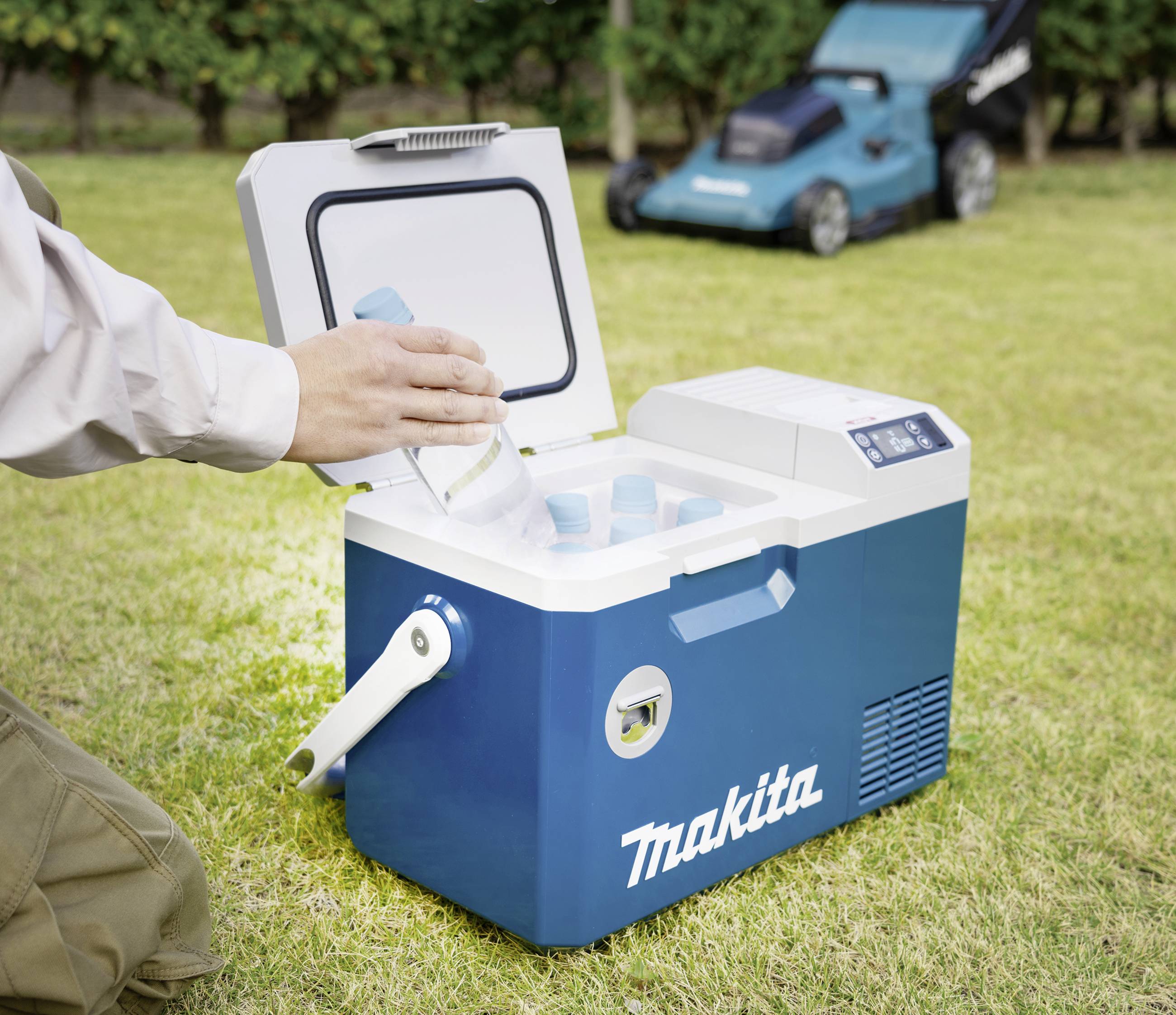 Een persoon legt een fles in een blauwe Makita-koelbox op een grasveld. Op de achtergrond staat een grasmaaier.
