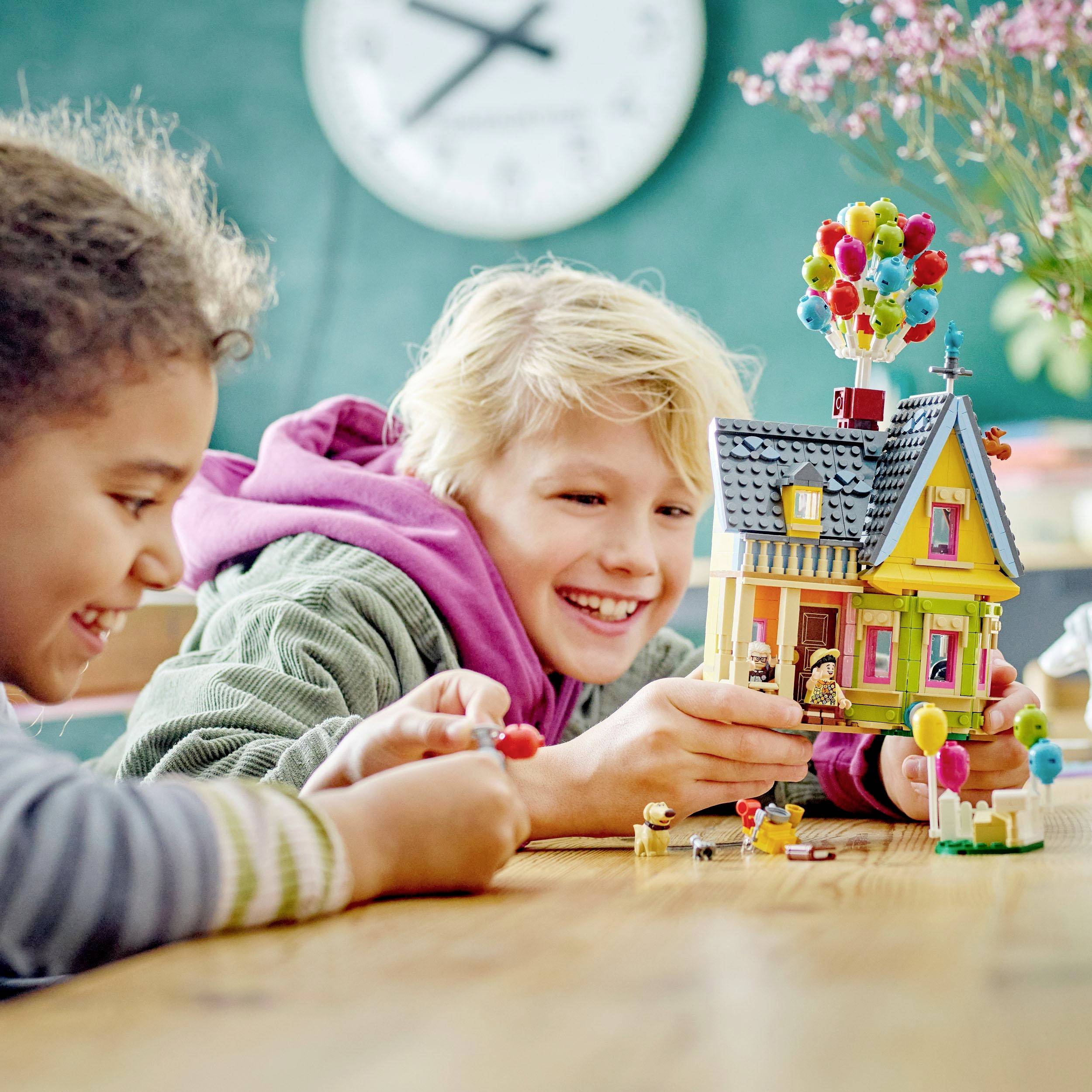 Twee kinderen spelen met een kleurrijk modelhuis van bouwstenen, dat wordt gedragen door luchtballonnen. Op de achtergrond is een klok zichtbaar.