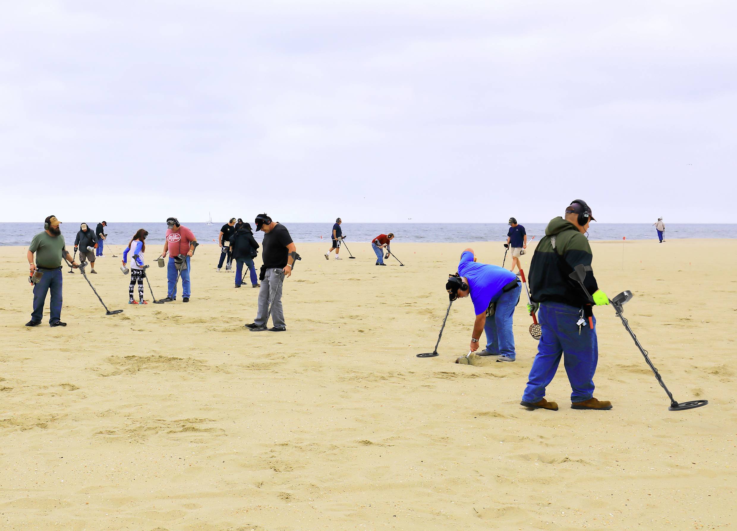 Mensen met metaaldetectoren doorzoeken een zandstrand met uitzicht op zee. Het is bewolkt, en het strand is nauwelijks bezet.