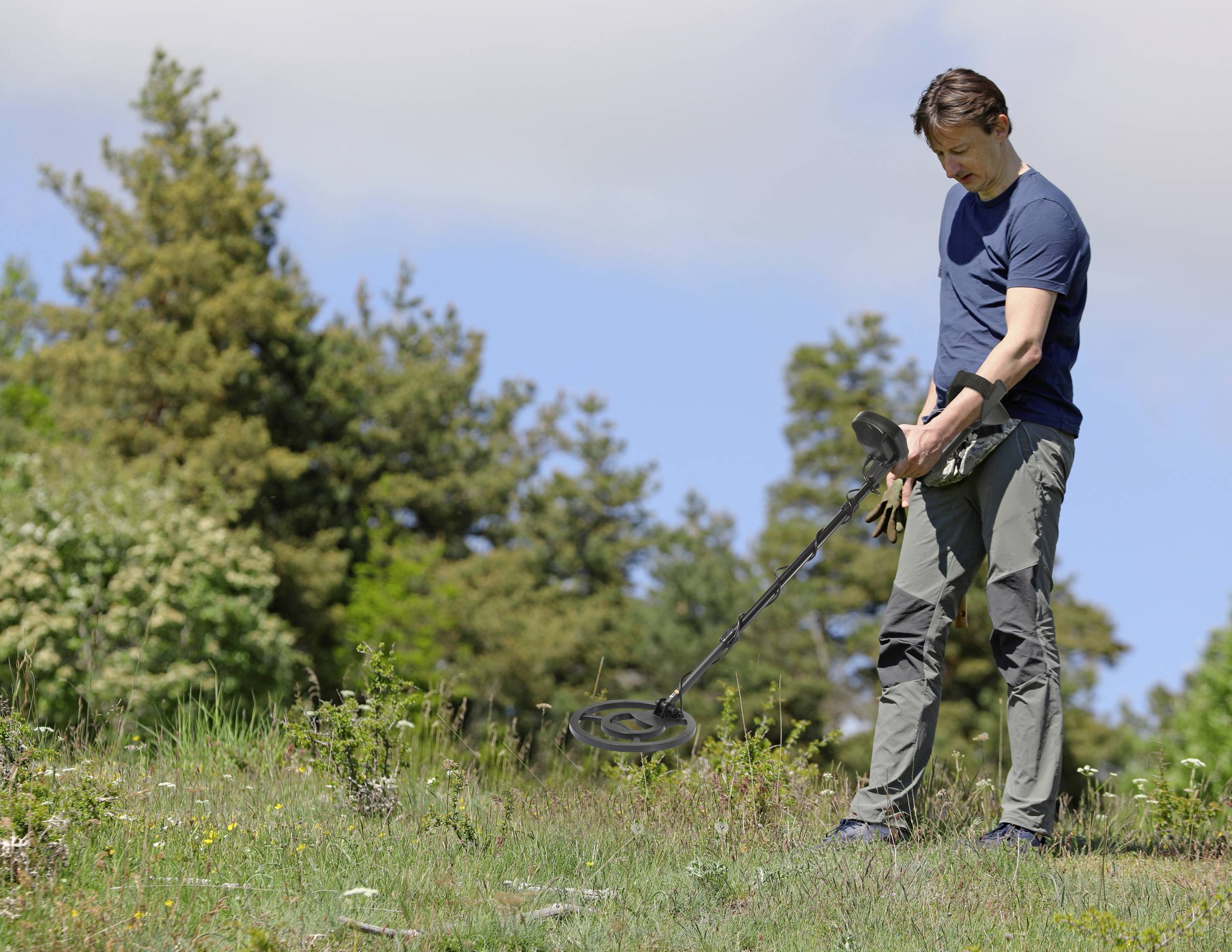 Een man gebruikt een metaaldetector op een weide met bomen op de achtergrond. Hij concentreert zich op de grond.