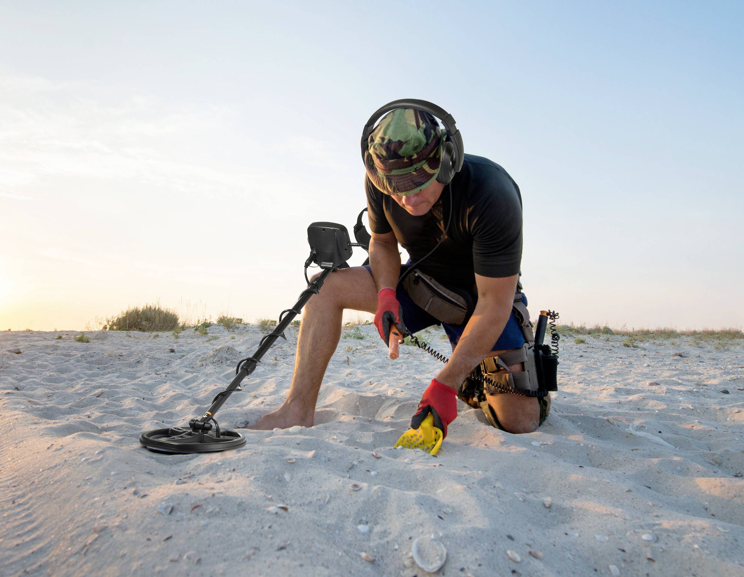 Persoon met metaaldetector knielt op het strand en onderzoekt het zand. Het is zonnig. Op de achtergrond zijn grassen en een blauwe lucht te zien.
