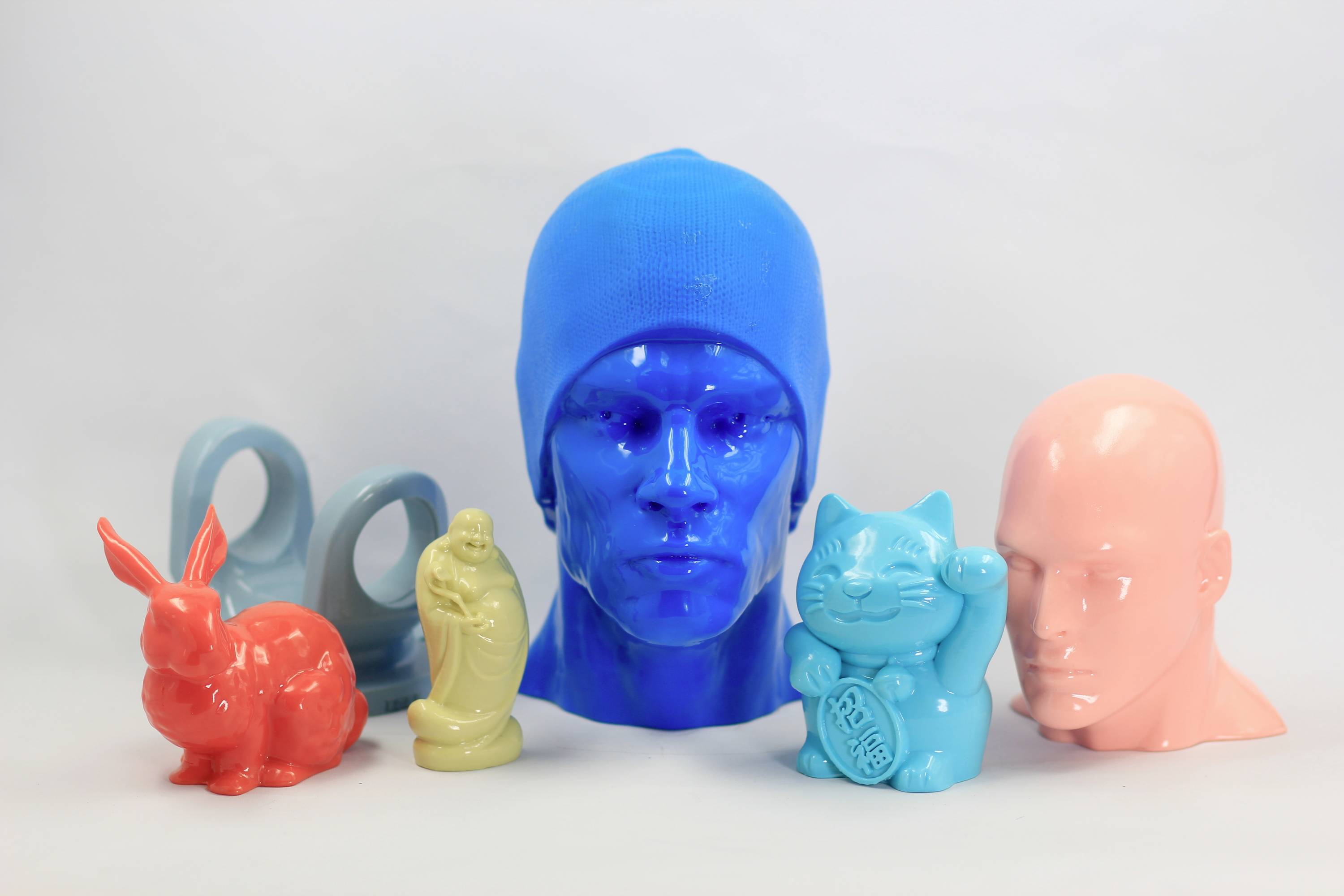 Gekleurde 3D-geprinte figuren: een blauwe kop met muts, een roze kop, een rode haas, een beige figuur, een lachende blauwe kat en twee blauwe ringen.