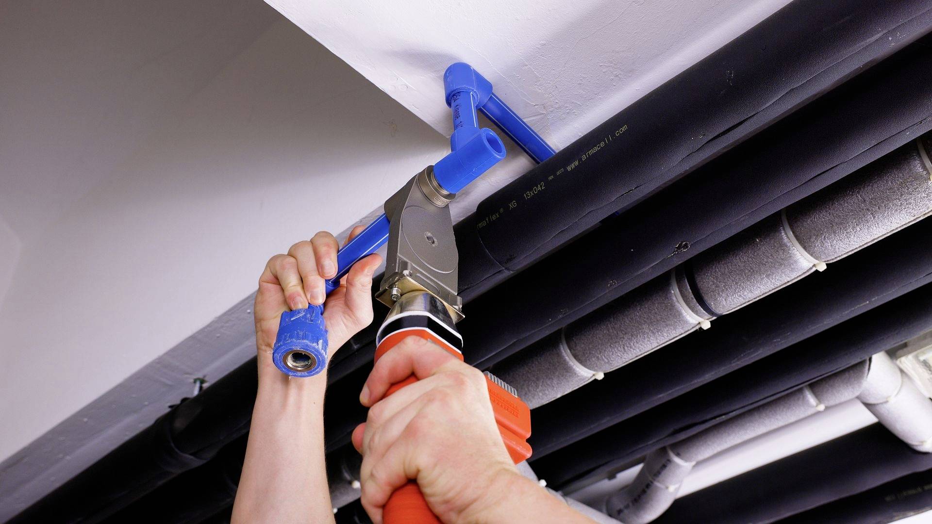 Een persoon installeert een blauwe waterleiding aan het plafond met een modern persgereedschap. Buizen lopen parallel langs het plafond.