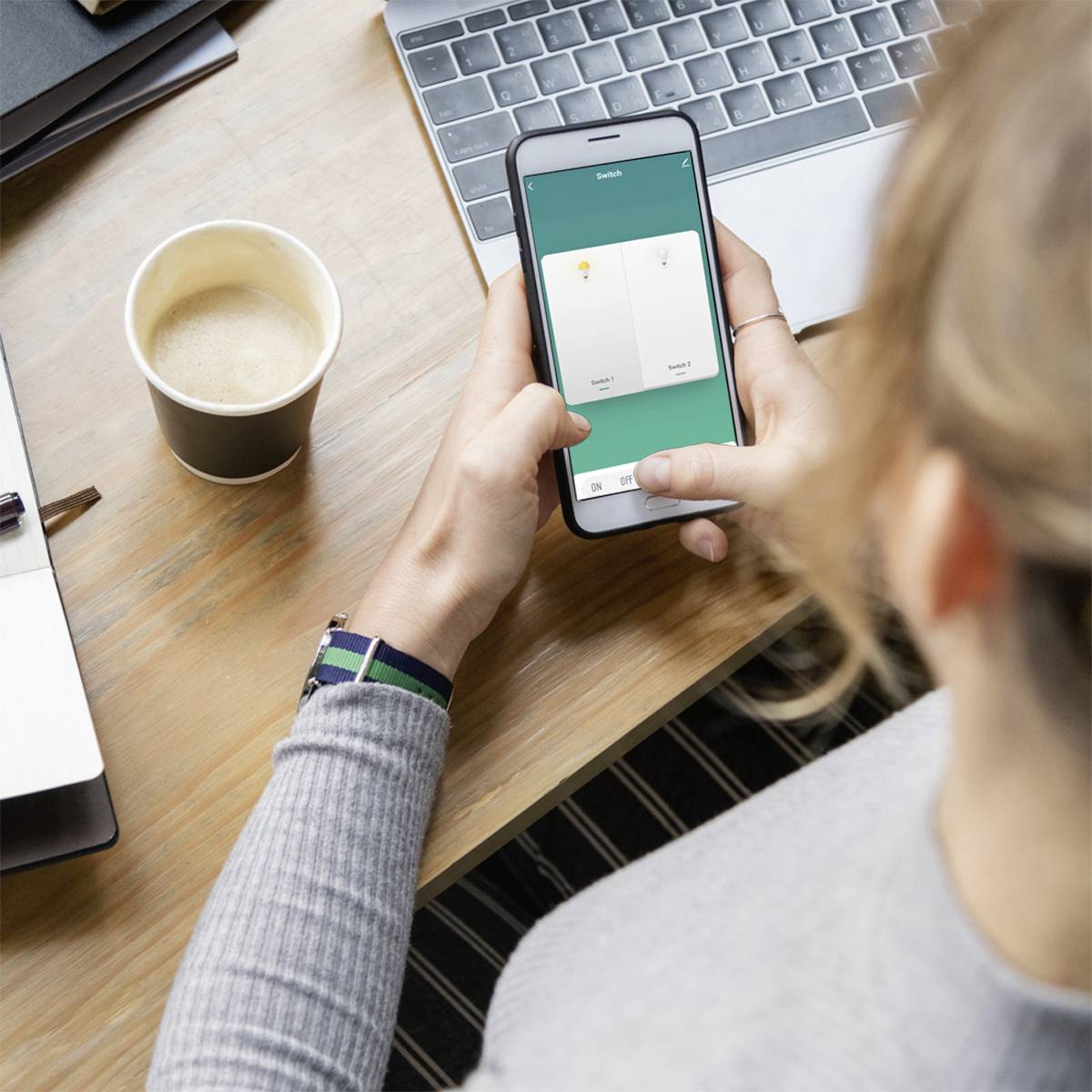 Een persoon bekijkt een app op een smartphone, terwijl een kopje koffie en een notitieboekje op tafel liggen.