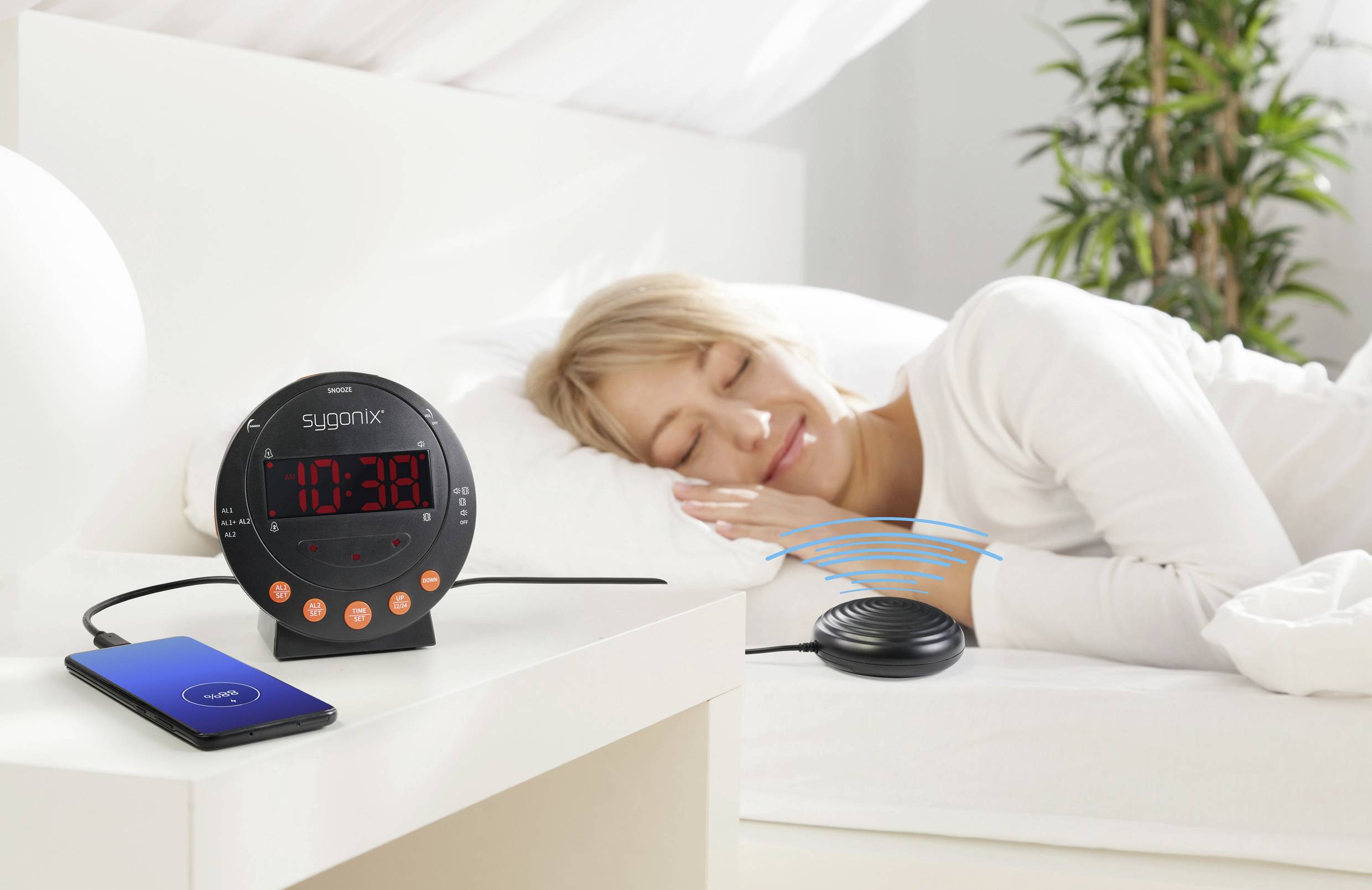 Een vrouw slaapt in bed. Naast haar staat een wekker met een grote LED-display. Een mobiele telefoon wordt opgeladen op het nachtkastje.