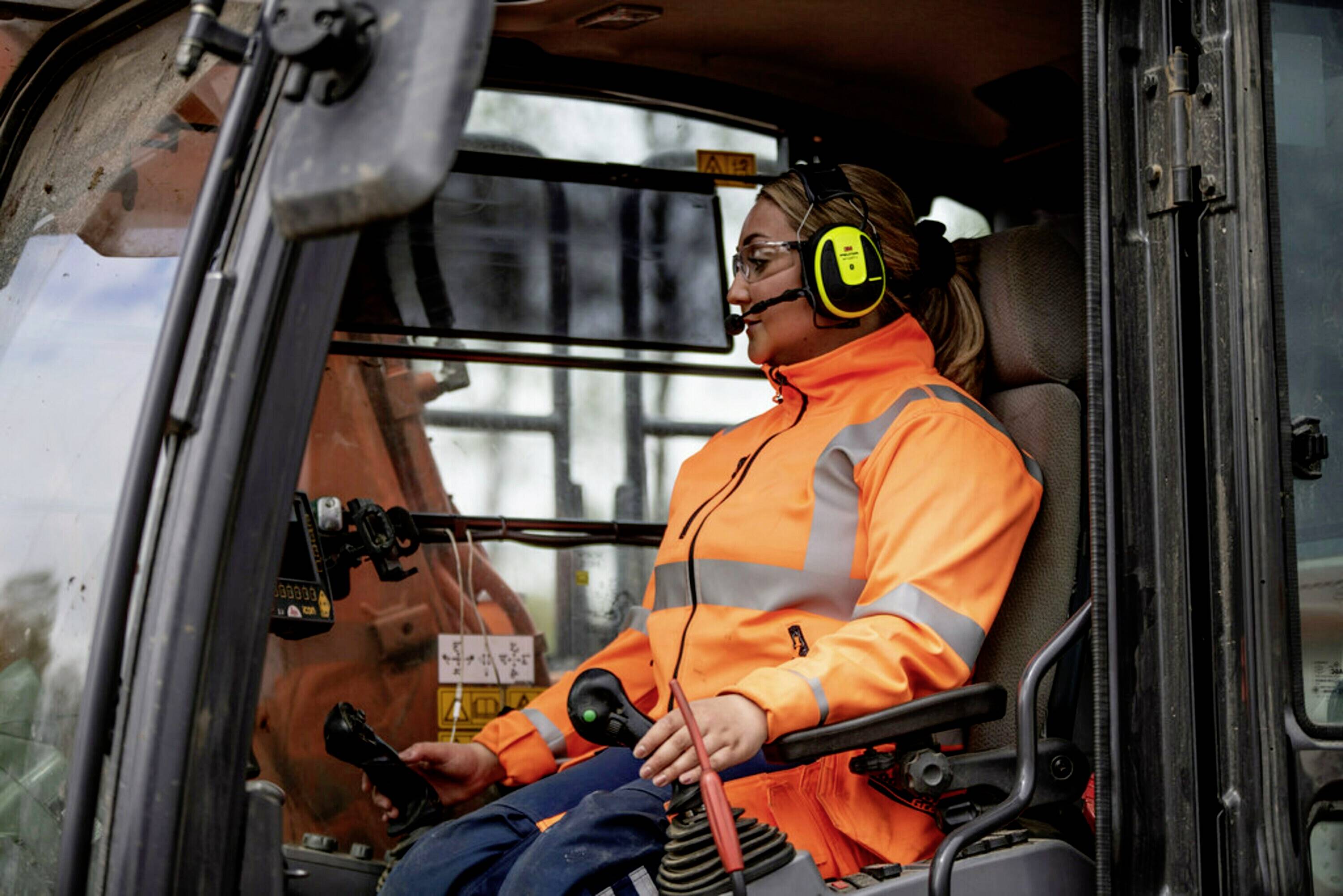 Een persoon in oranje waarschuwingskleding bedient de stuurhendels binnenin de bestuurdercabine van een bouwmachine.