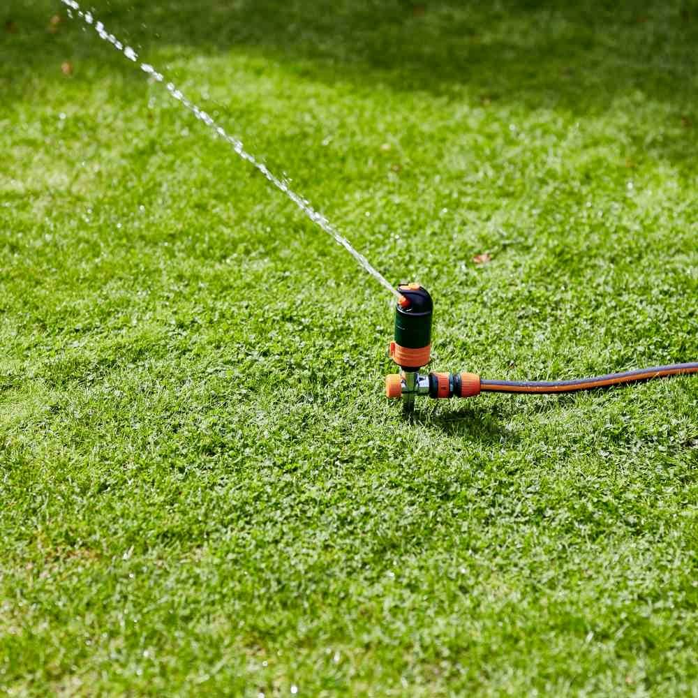Een sproeier met een oranje aansluiting besproeit het groene gras op een grasveld. De waterstraal verspreidt zich gelijkmatig.