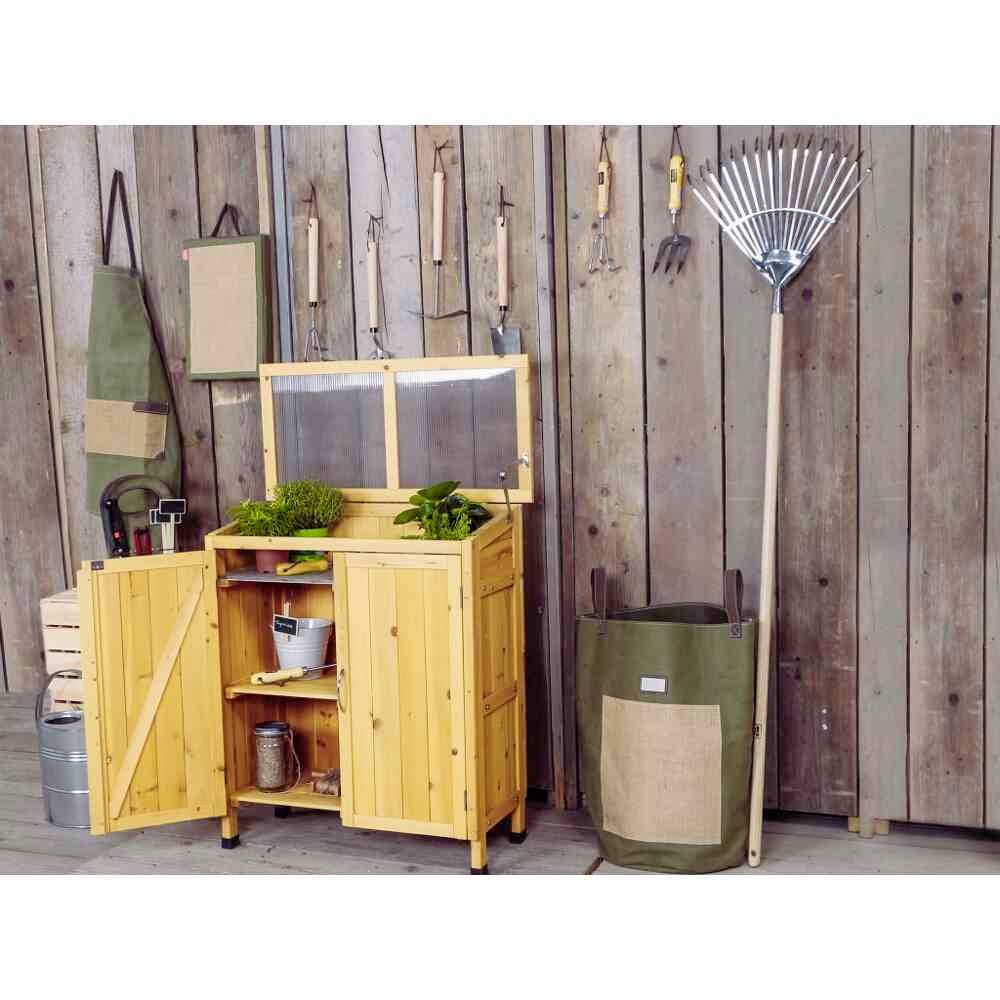 Een houten tuinkast staat open en toont planten en gereedschappen. Aan de muur hangen meer tuingereedschappen en een schort.