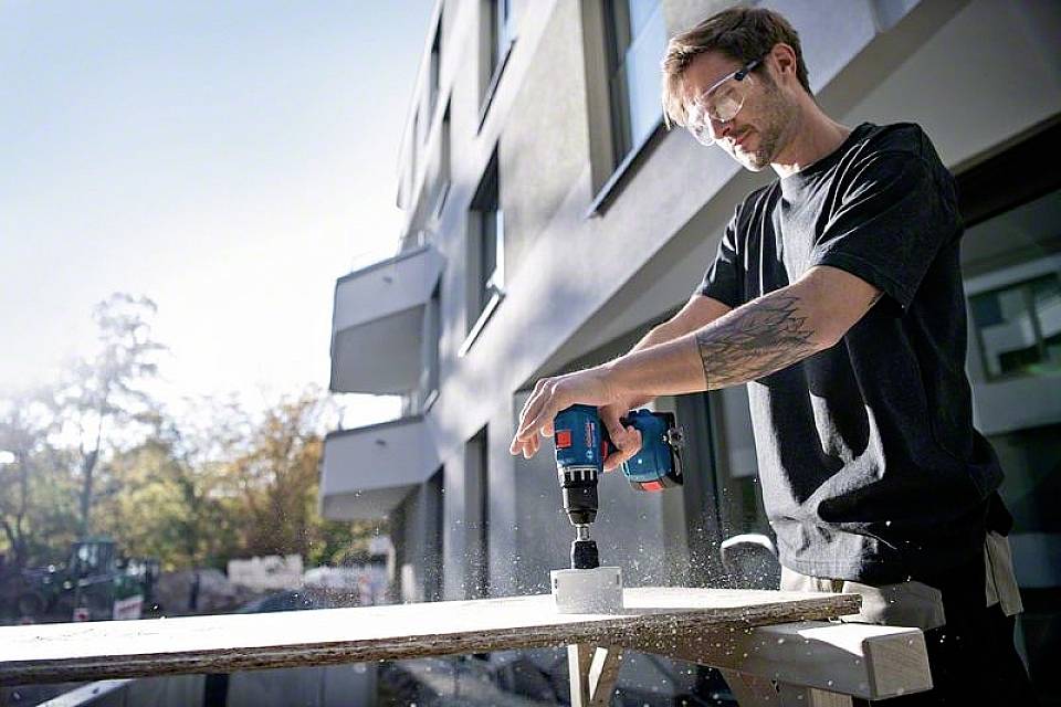Een man boort met een elektrische boormachine een gat in een houten plank op een bouwplaats, draagt een veiligheidsbril.
