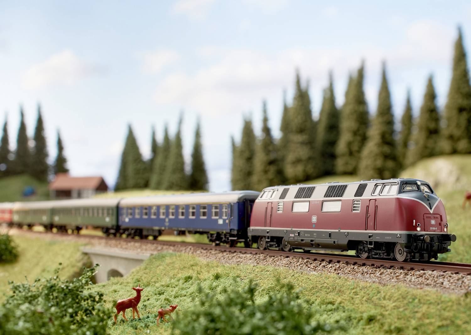 Modelspoorbaan landschap met trein: Een rode trein rijdt over een spoor door een groene omgeving met dennenbomen en twee reeën op de voorgrond.