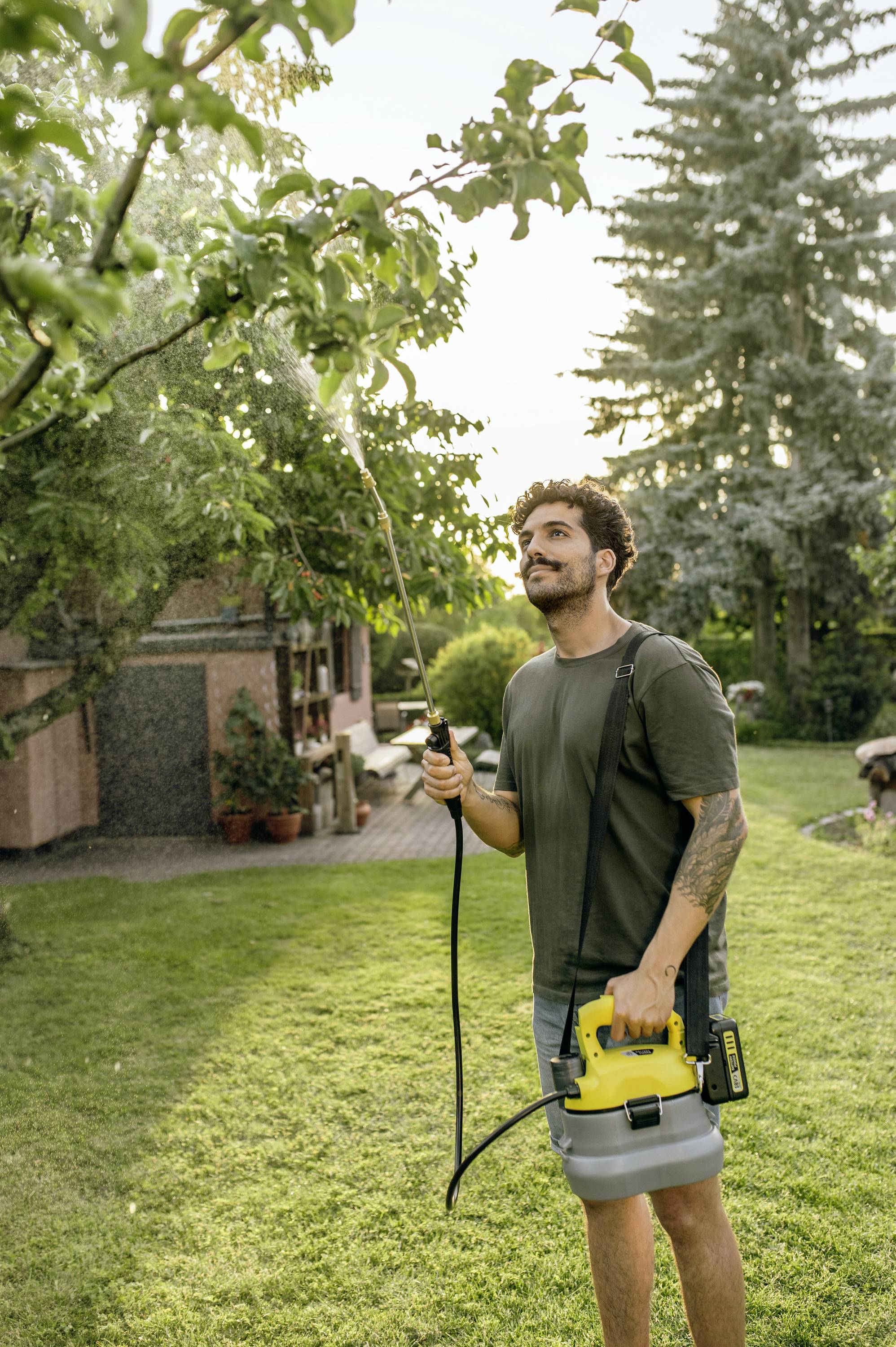 Een man besproeit een boom in de tuin met een handmatig apparaat. Op de achtergrond zijn huizen en bomen zichtbaar.