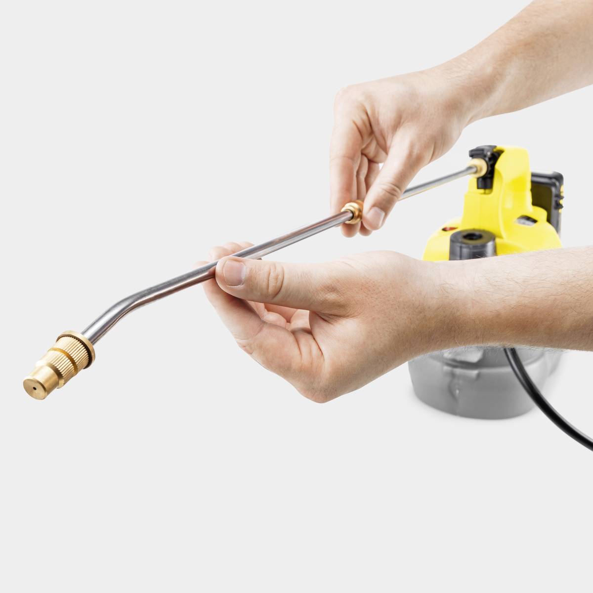 Een hand houdt een metalen buis vast die is aangesloten op een hogedrukreiniger. Op de achtergrond is een gele hogedrukreiniger zichtbaar.