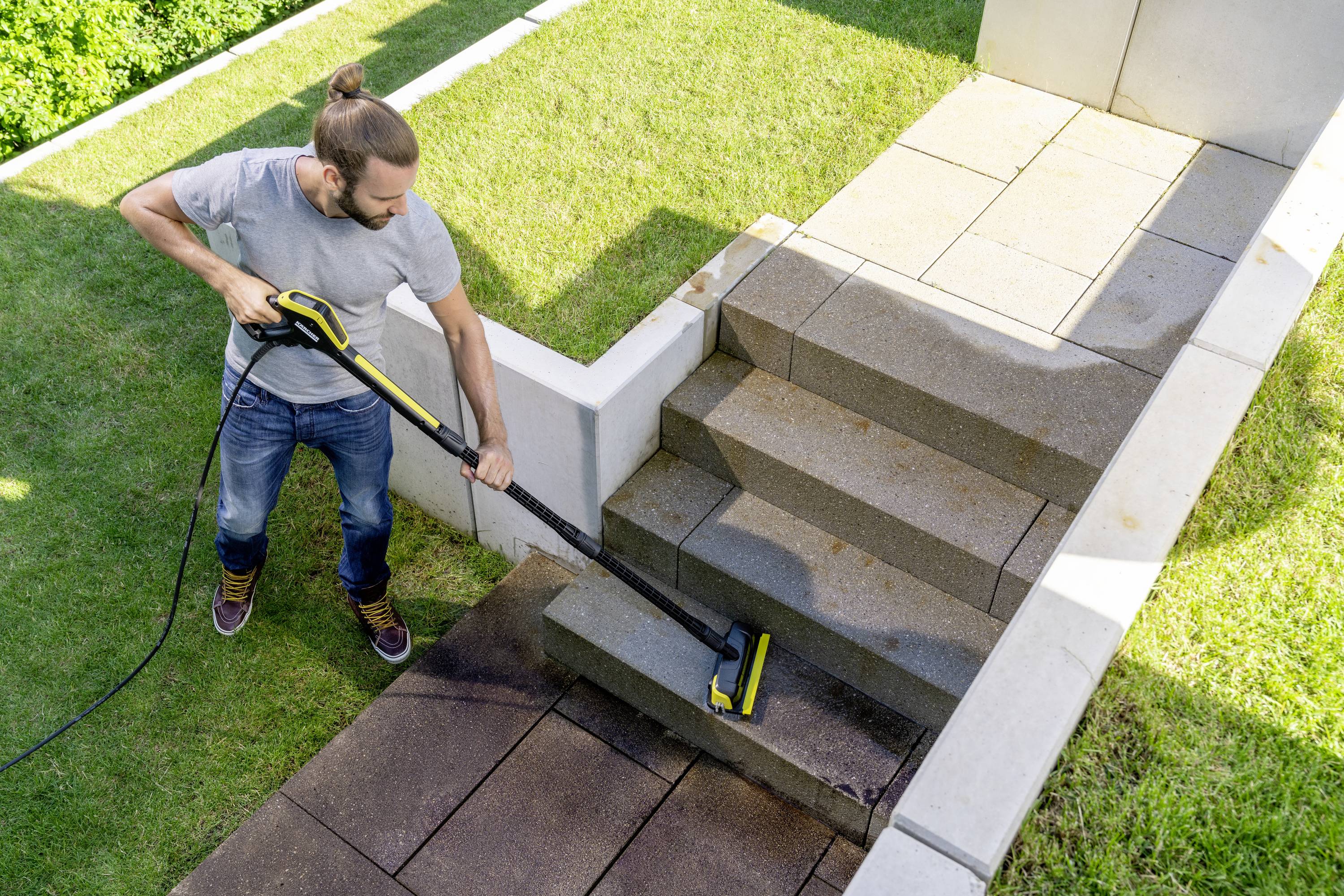 Een man reinigt stenen trappen op een terras met een hogedrukreiniger. Om hem heen zijn grasvelden zichtbaar.