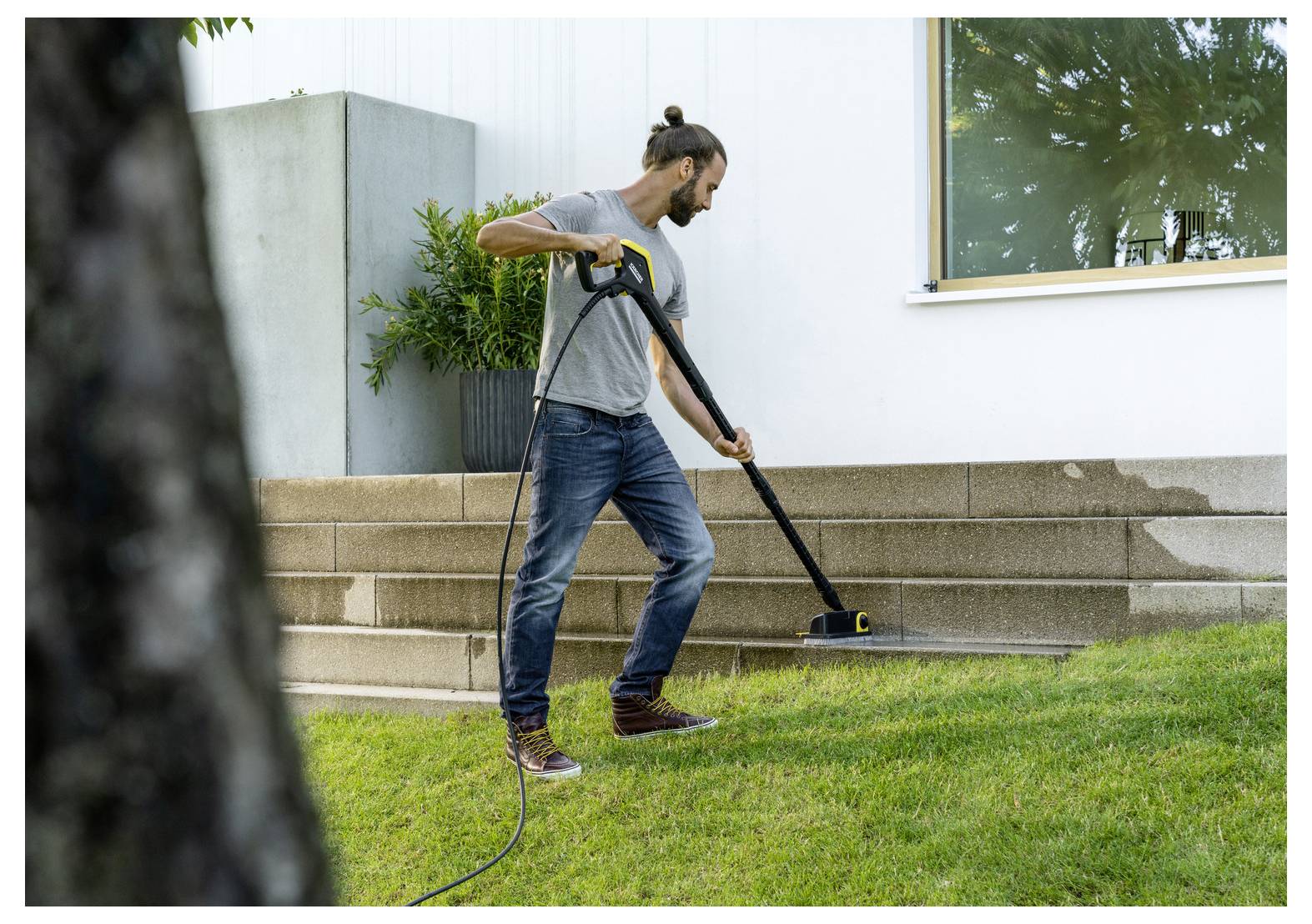 Iemand reinigt buitentrappen met een hogedrukreiniger, waardoor vuil wordt verwijderd van het stenen oppervlak. Een pot met planten is zichtbaar in de buurt.
