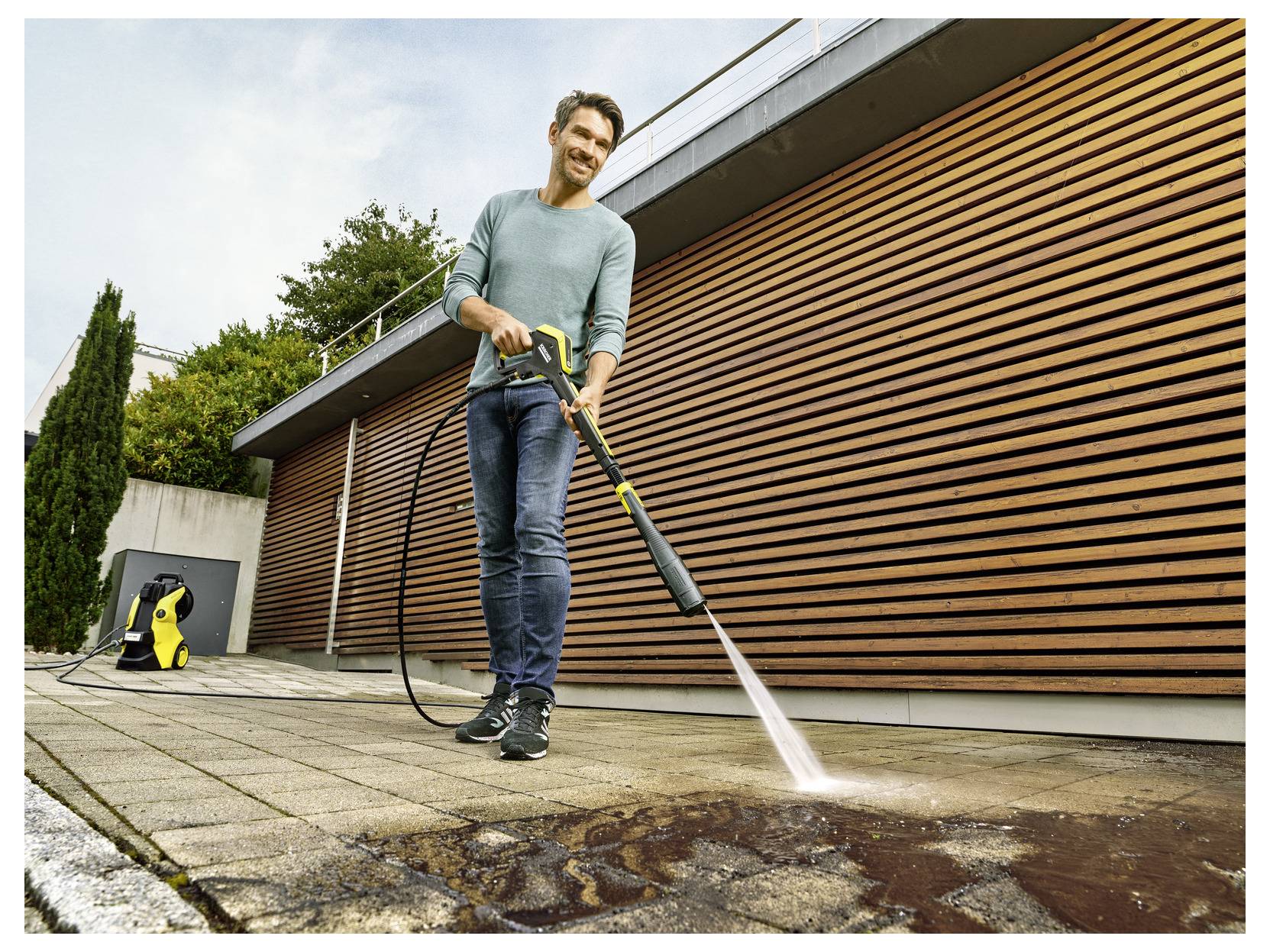 Een persoon reinigt een buitenpatio met een hogedrukreiniger, waarbij vuil wordt verwijderd. Hij/zij gebruikt een gele hogedrukreiniger die is aangesloten op een waterslang.