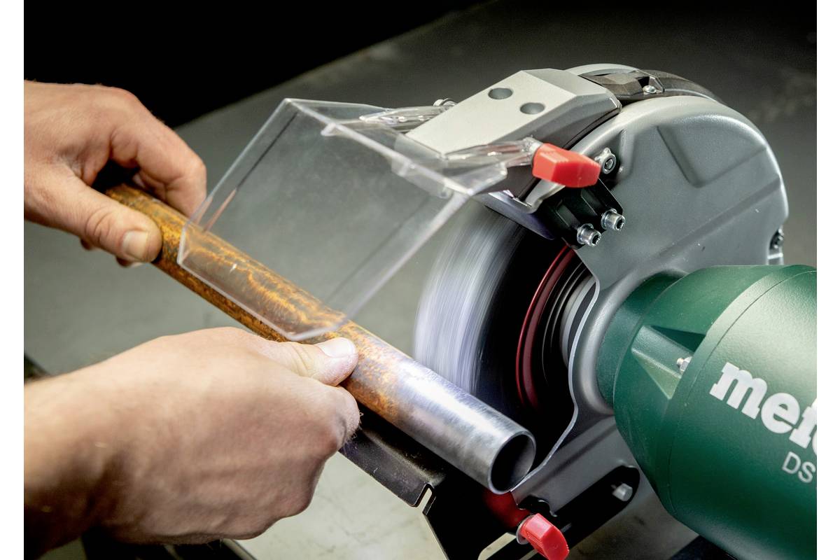Een persoon houdt een roestig pijp aan een slijpmachine. Er vliegen geen vonken, veiligheidsvoorziening aanwezig. Metabo-logo zichtbaar.