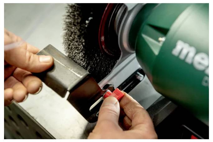 Handen die een machine met een staalborstel gebruiken om een metalen object te polijsten of schoon te maken. Het gereedschap heeft een groene behuizing en de machine is in werking.