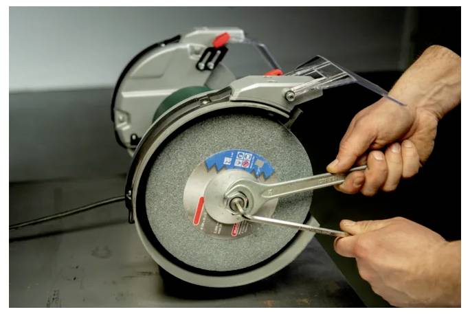 Persoon die een bankslipschijf afstelt met een kompas in de buurt van beschermende schermen, wat werktuigonderhoud en veiligheidsuitrusting toont.