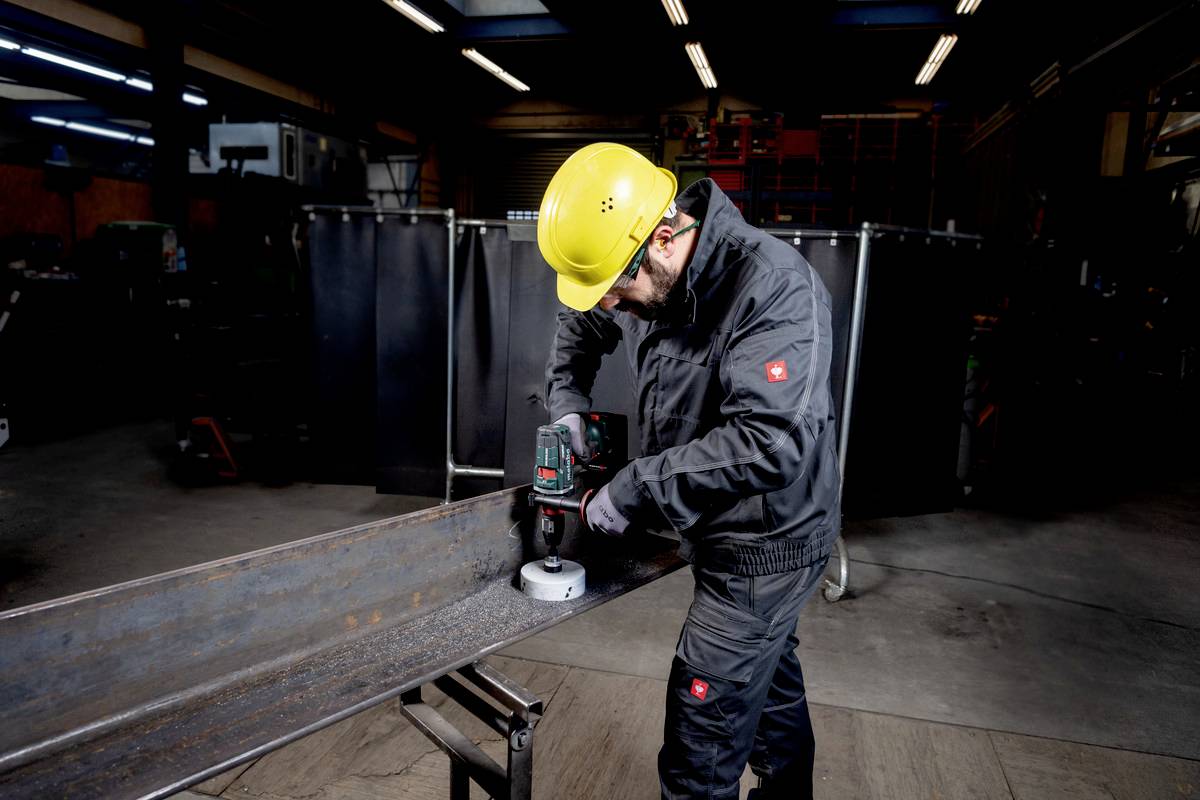 Een arbeider in beschermende kleding en helm boort met een machine in een stalen balk in een fabriekromgeving.