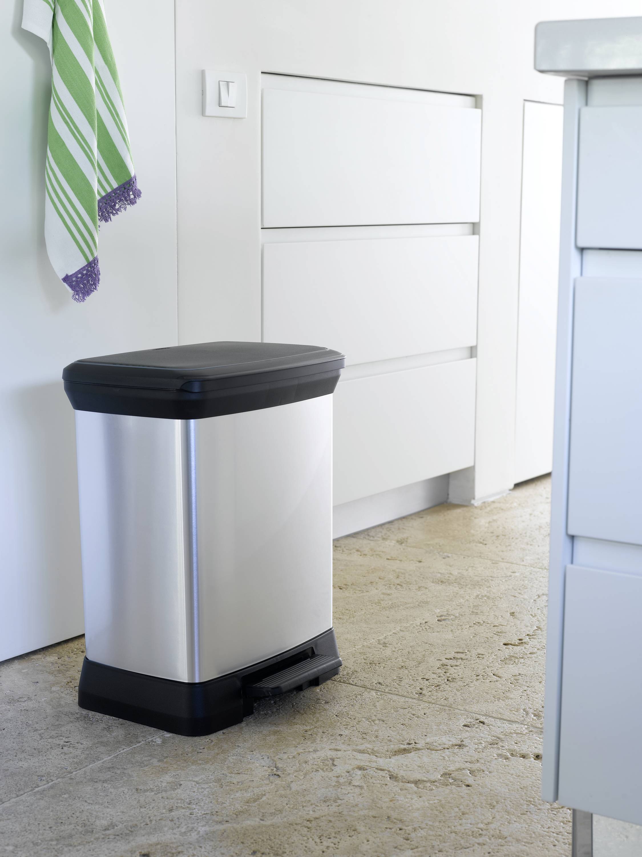 Metalen emmer met zwarte deksel en pedaal staat op stenen vloer in moderne keuken. Rechts hangen handdoeken.