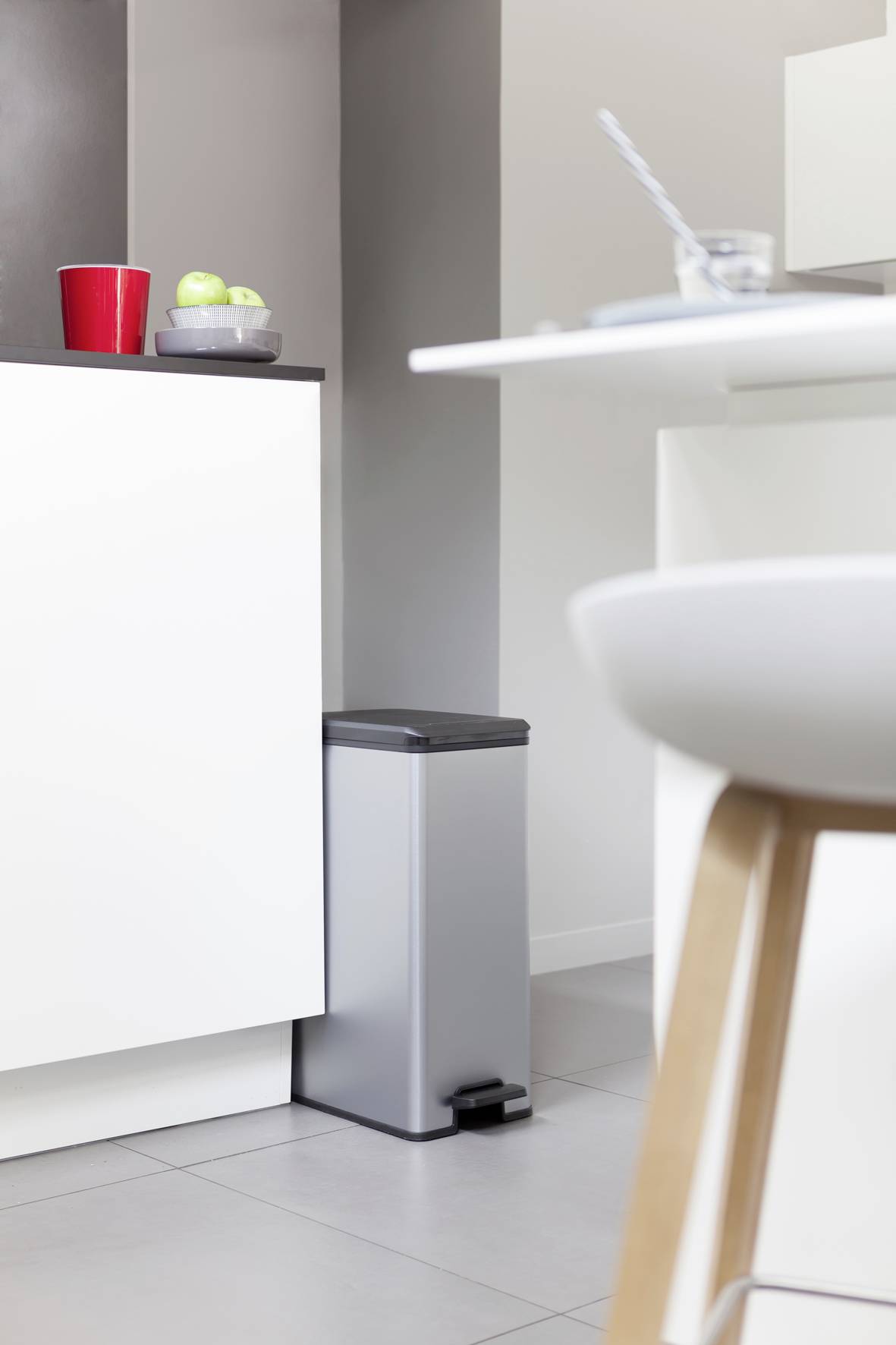 Grijze prullenbak met voetpedaal in moderne keuken. Links een aanrecht met rode mok en groene appels.