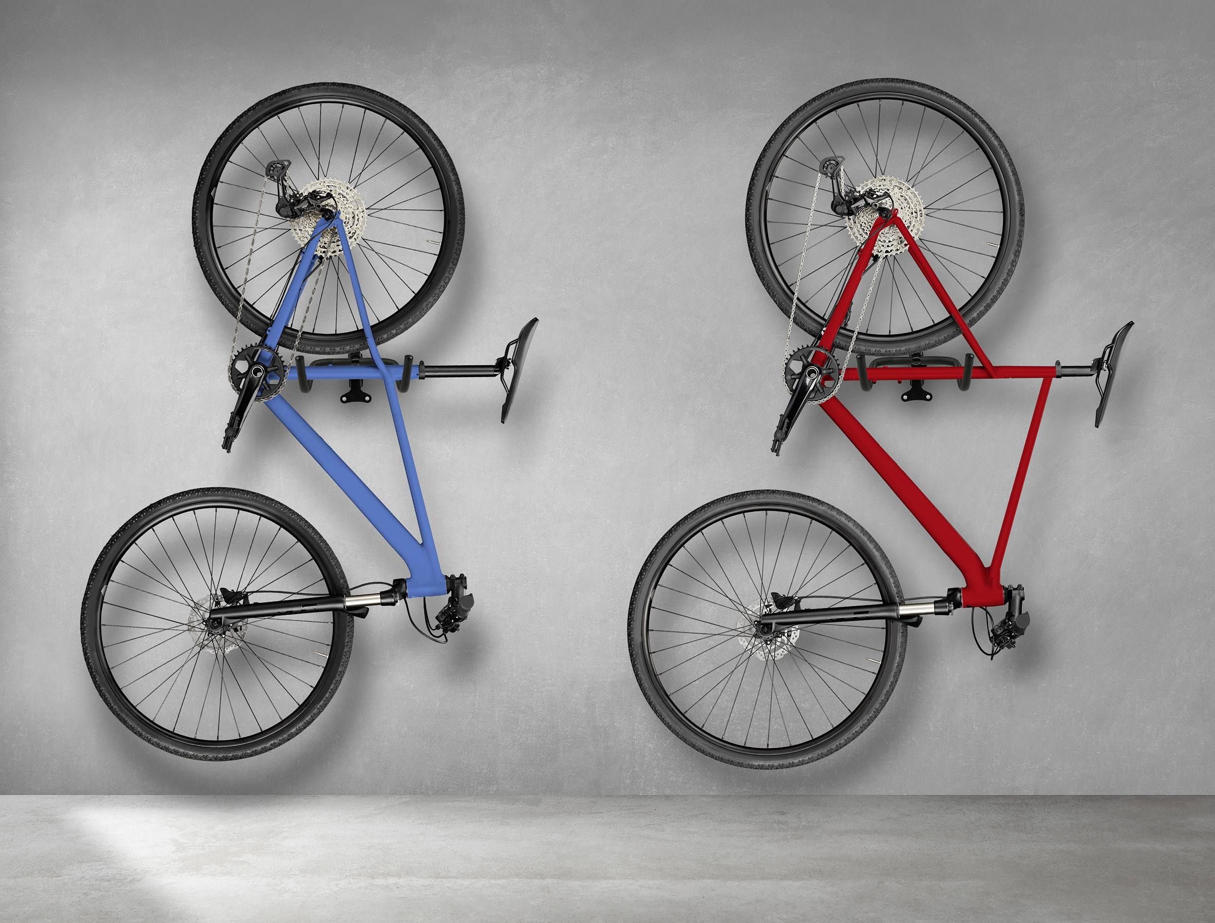 'Twee fietsen, een blauwe en een rode, hangen verticaal aan een grijze muur in een moderne tentoonstellingsruimte.'