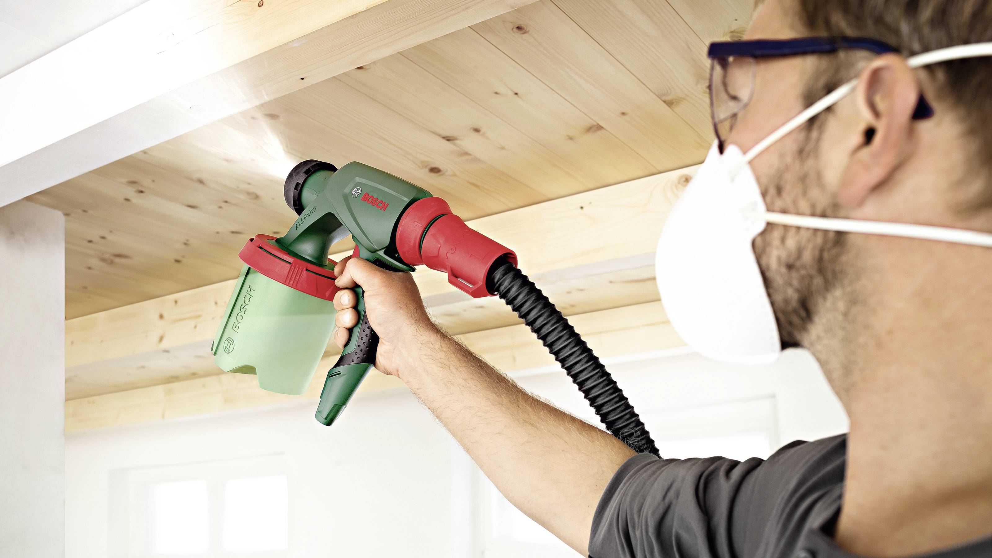 Een persoon slijpt een houten plafond met een elektrisch gereedschap. Hij draagt een veiligheidsbril en een stofmasker.