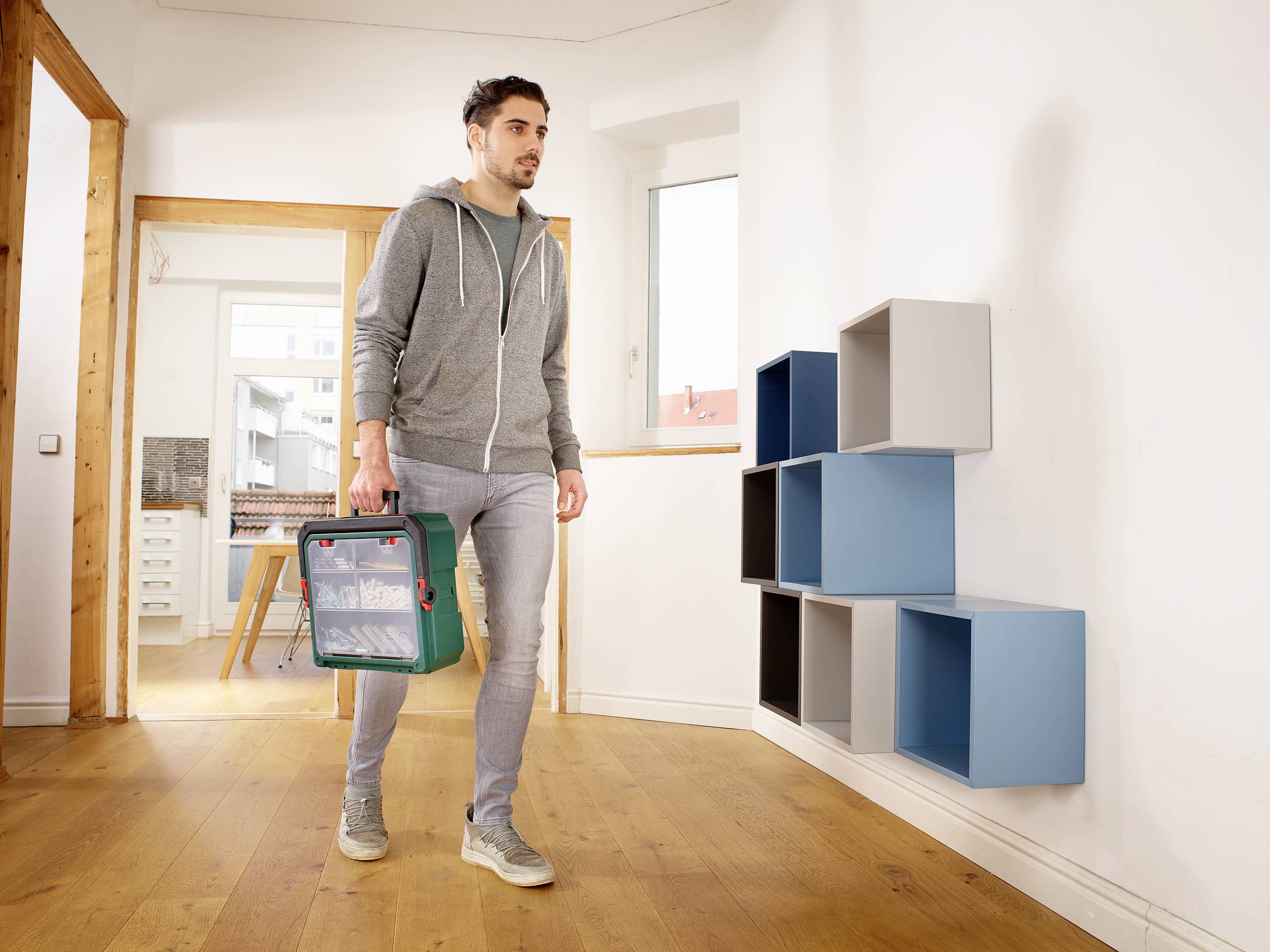 Een man in vrijetijdskleding loopt door een ruimte met een gereedschapskist. Aan de muur zijn gekleurde opbergkubussen bevestigd.