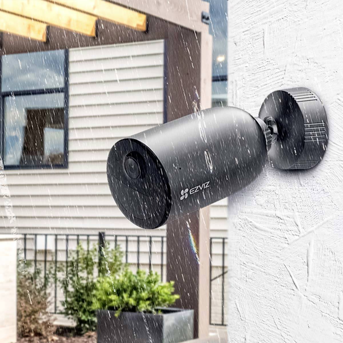 Bewakingscamera aan een muur, zichtbaar in de regen, kijkt uit op een modern huis met houten bekleding en grote ramen op de achtergrond.