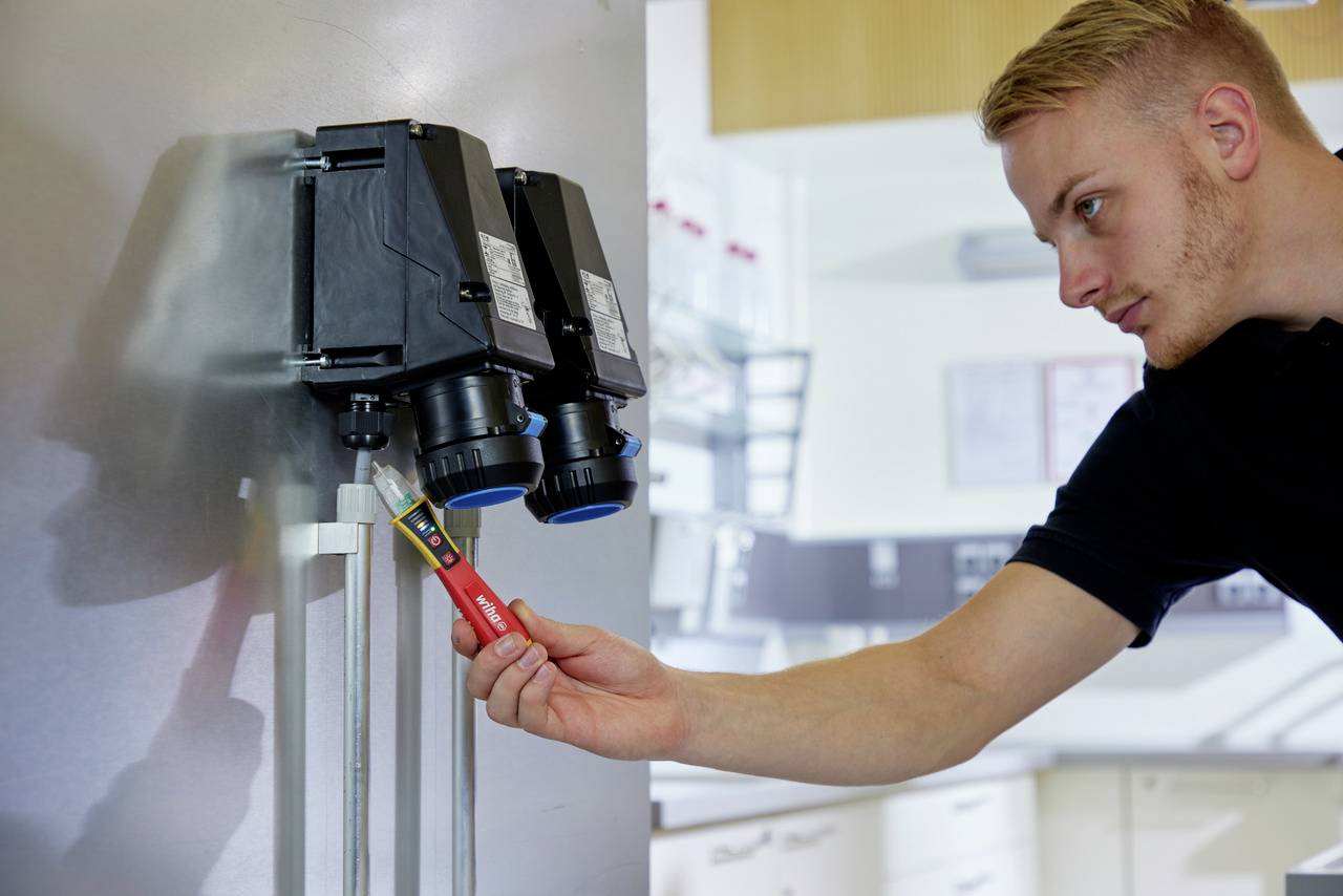 Een man controleert de elektrische aansluitingen in een schakelkast met behulp van een spanningszoeker. De achtergrond toont een laboratoriumomgeving.