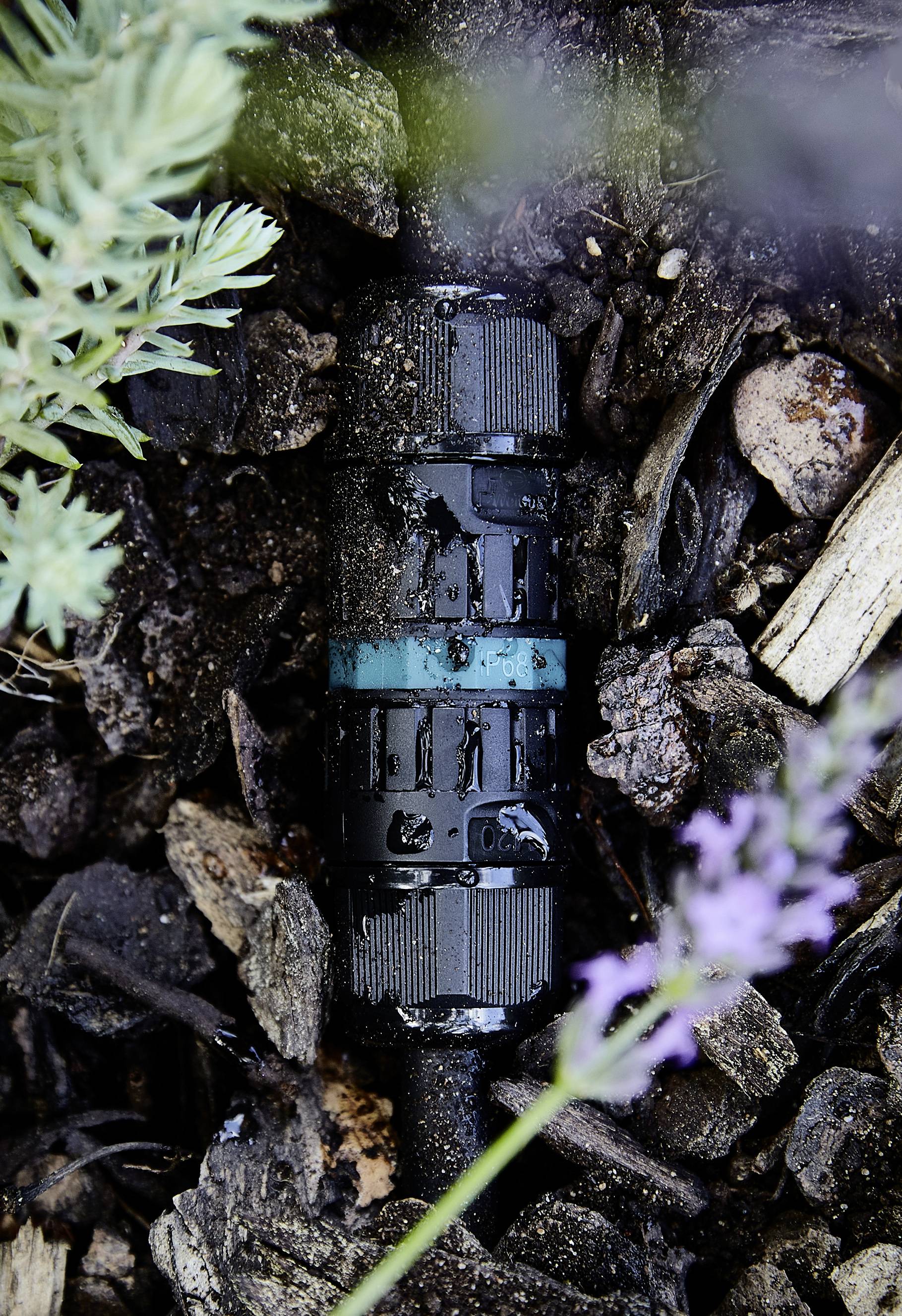Een zwarte tuinslangverbinder ligt op vochtige grond, omringd door planten en houten stukken, licht bevochtigd met water.