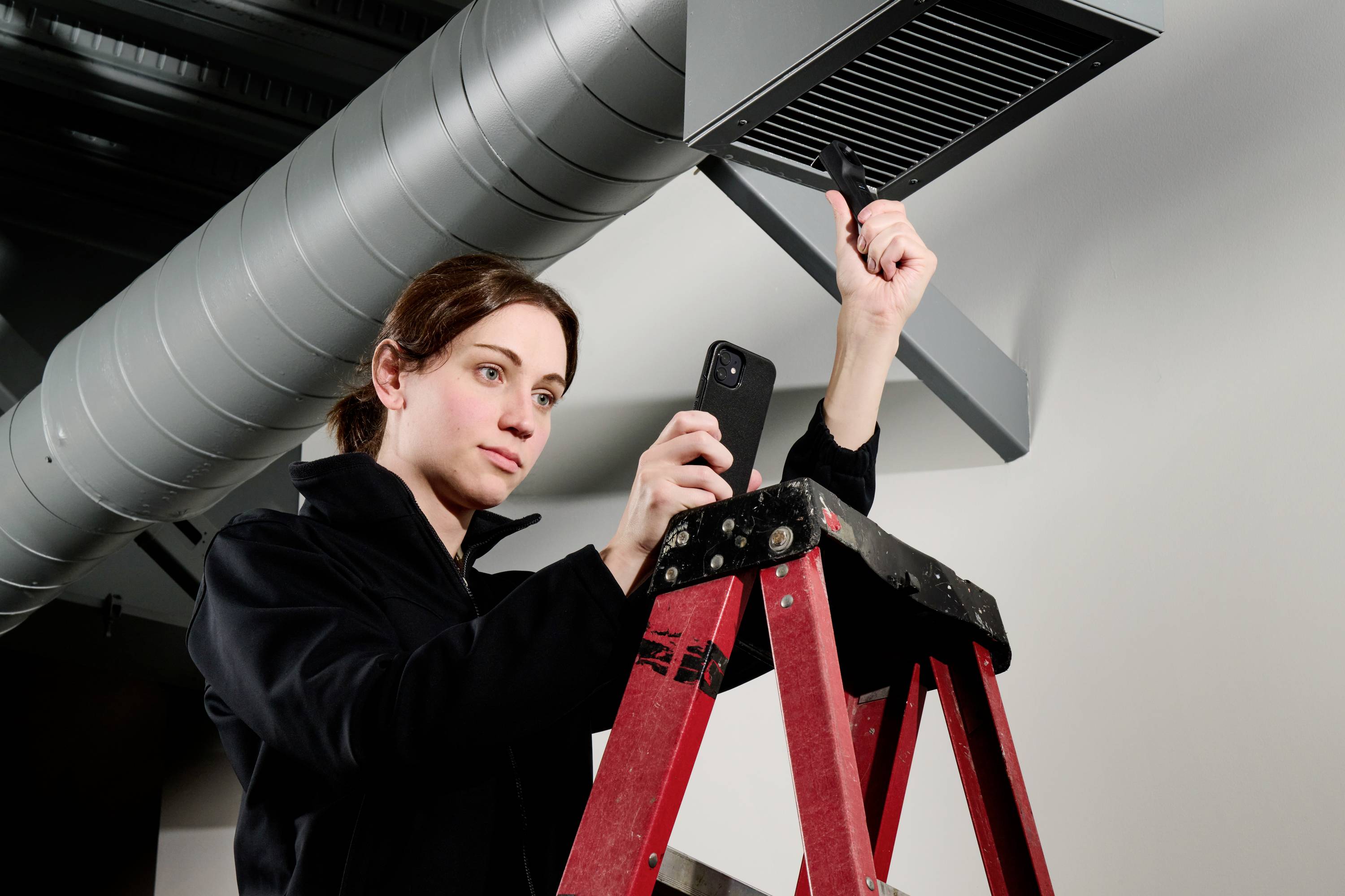 Een persoon op een ladder inspecteert een ventilatieschacht, terwijl hij een smartphone en een inspectietoestel vasthoudt.