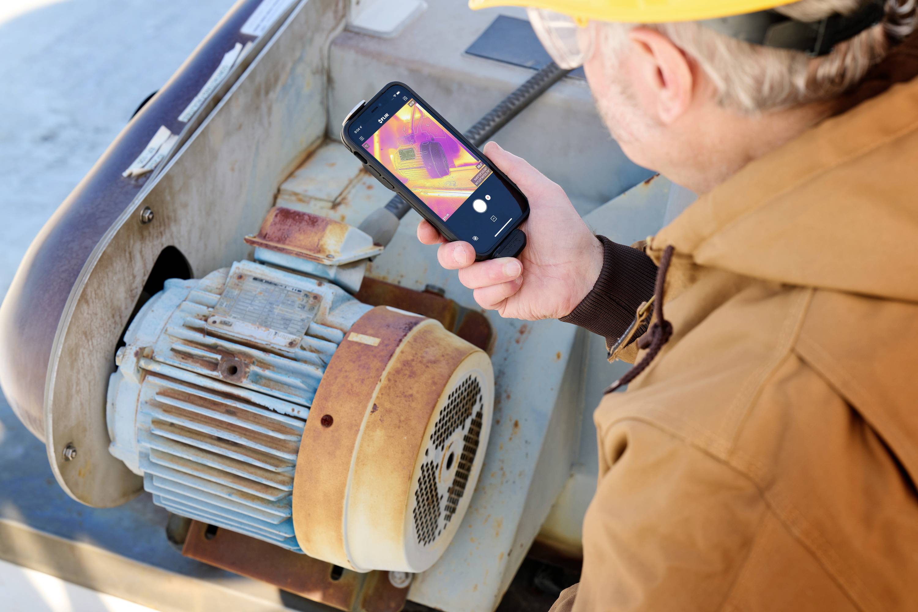 Een persoon met een gele veiligheidshelm inspecteert een motor en gebruikt een smartphone met warmtebeeldcamera om de temperatuur te controleren.