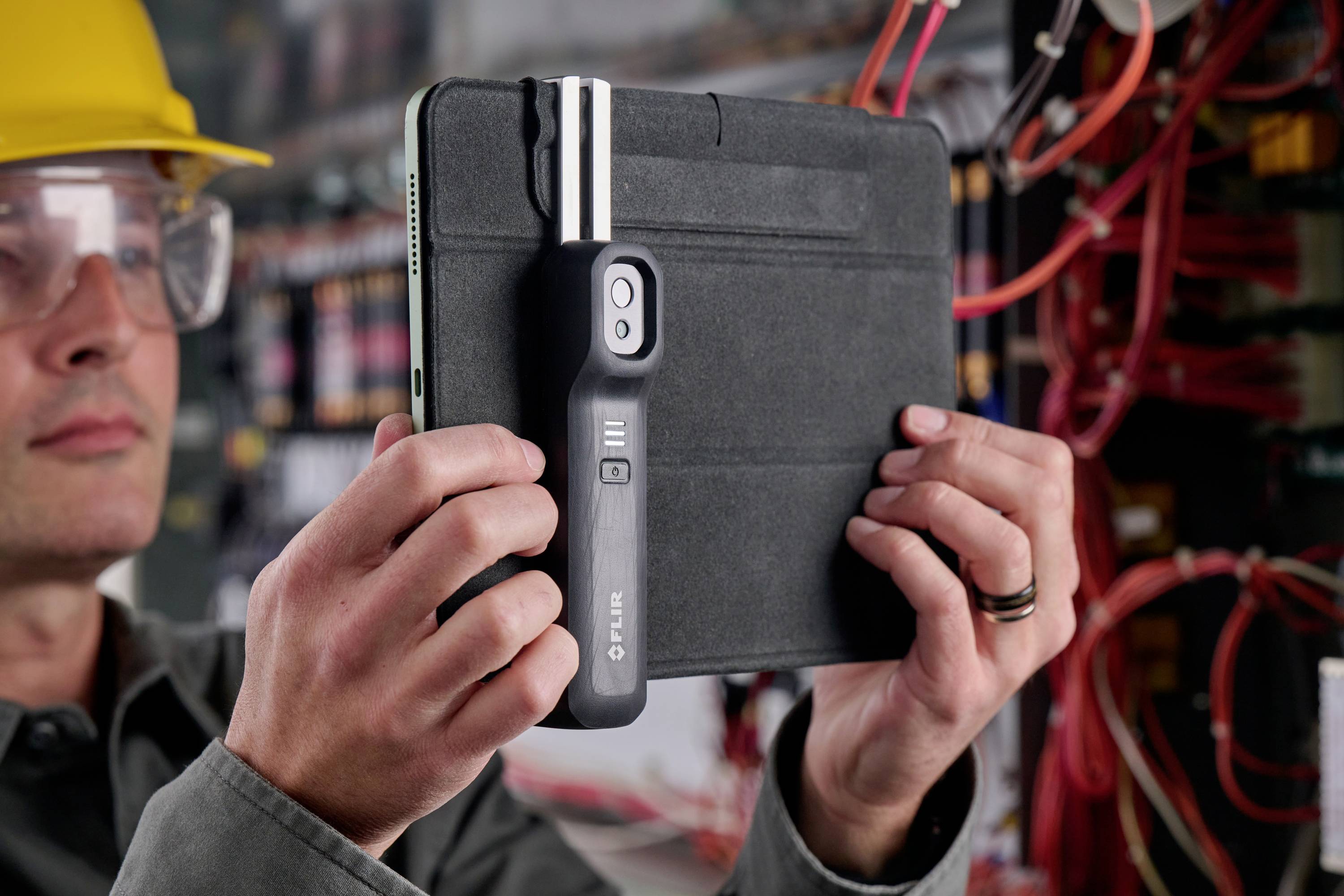 Een persoon houdt een tablet vast met een opzetstuk voor het meten van elektrische installaties, terwijl hij een kabelsysteem controleert.