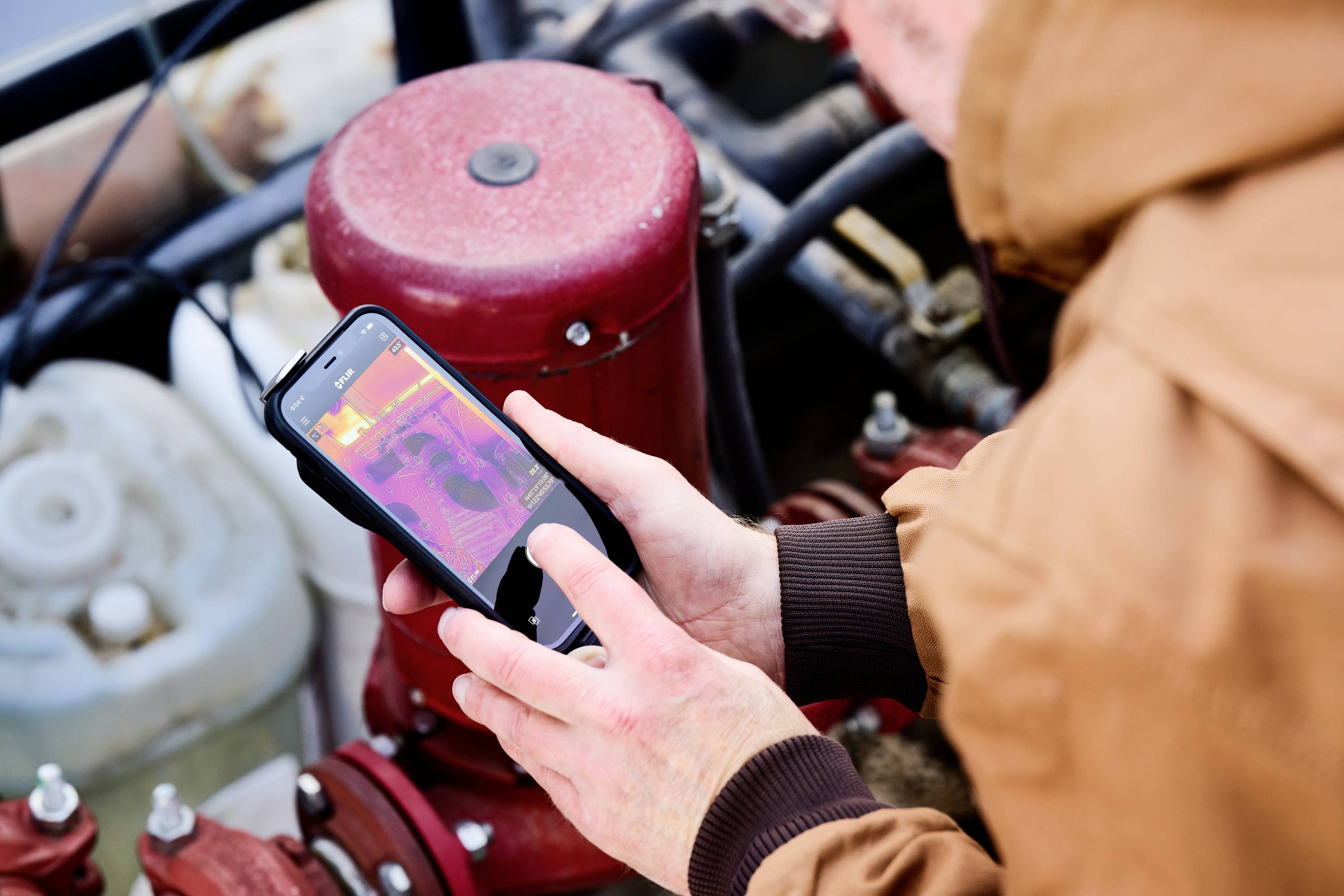 Persoon controleert verwarmingssysteem met smartphone en warmtebeeldapp. Rode, roze en witte kleuren tonen temperatuurbereiken.
