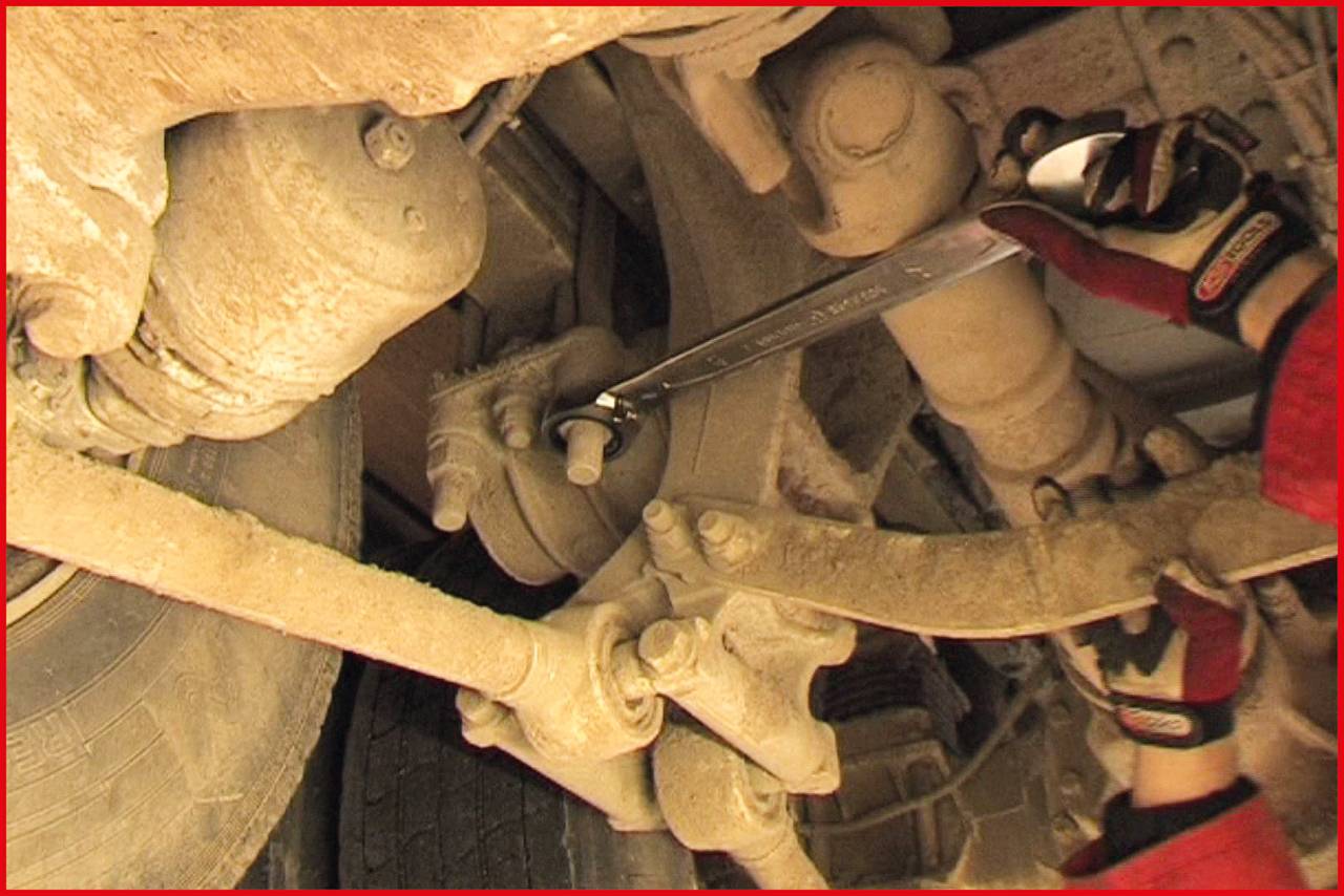 Een persoon repareert de onderkant van een voertuig met een moersleutel, gefocust op een as naast de band.